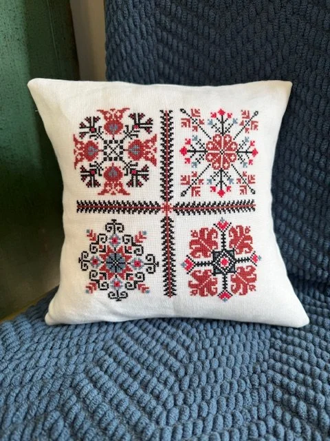 Decorative pillow with cross-stitch patterns in red, black, and gray on white fabric, divided into four sections by a cross, each with a unique symmetrical design.