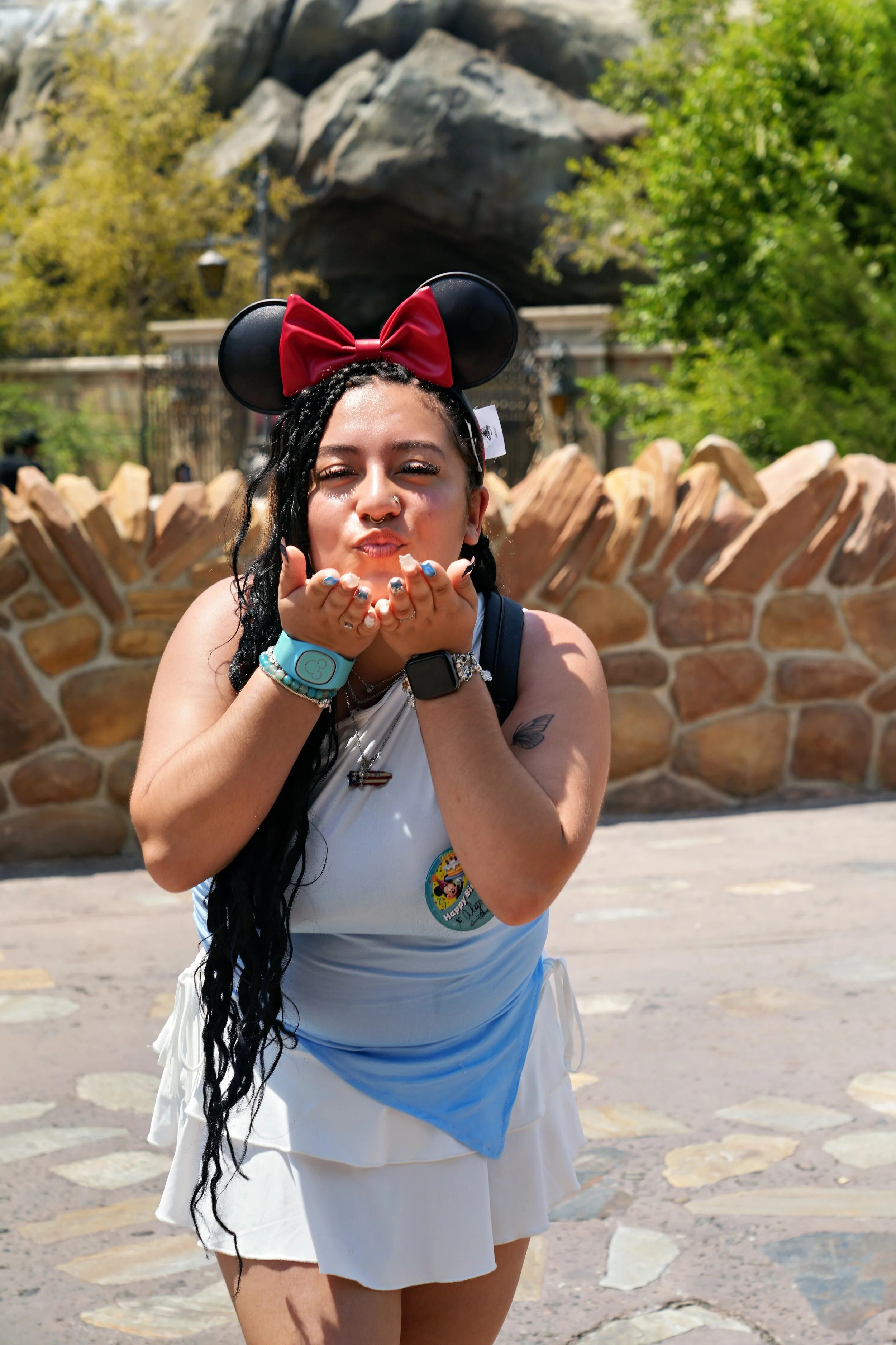 A woman with long black hair wearing a Minnie Mouse ears headband, blowing kisses, standing outdoors with a stone wall and greenery in the background.