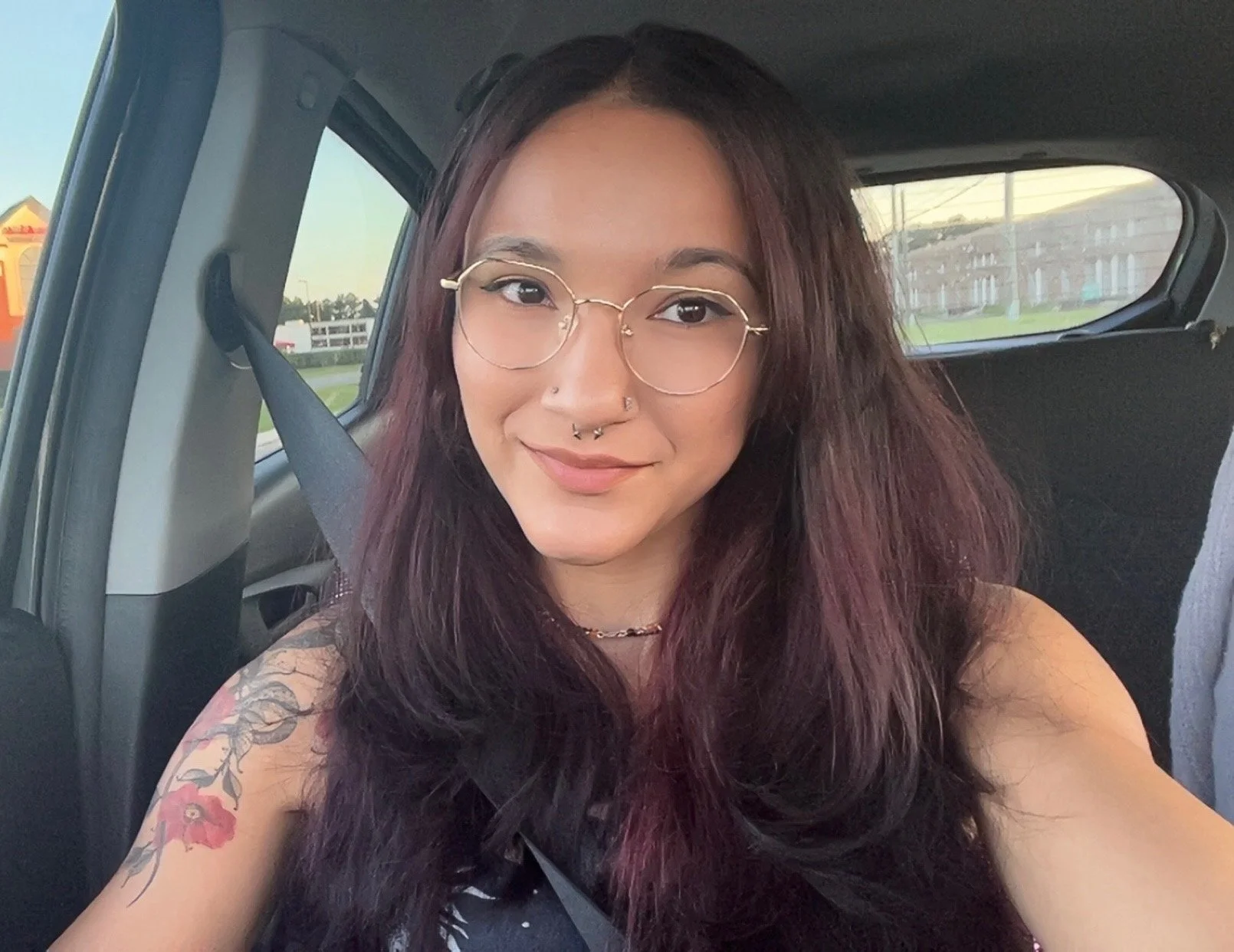 Young woman with long dark hair, glasses, and tattoos sitting in a car, smiling at the camera during daylight.
