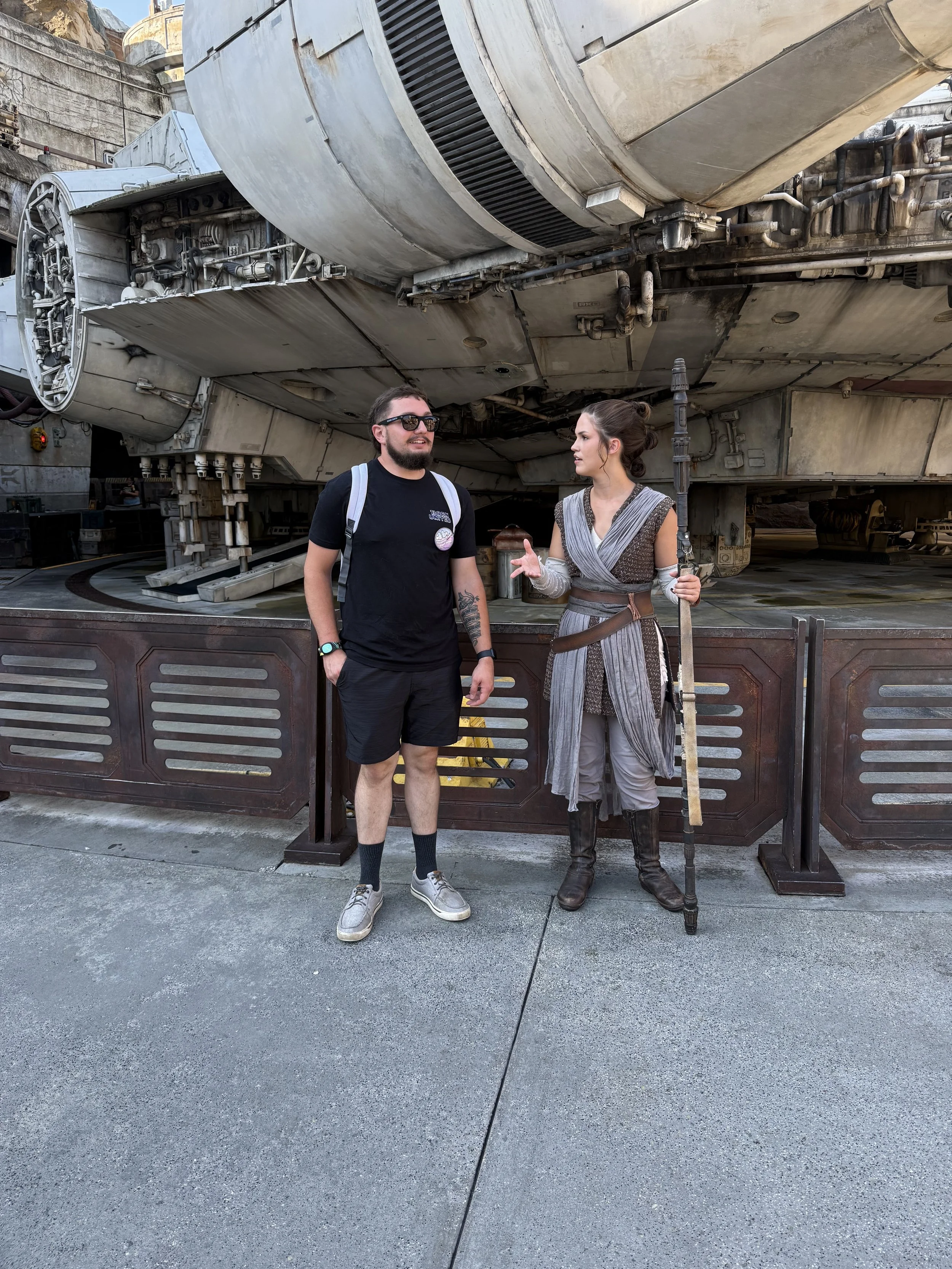 A man and a woman dressed as a Jedi from Star Wars are standing in front of the Millennium Falcon. The woman is holding a staff and gesturing while talking to the man.