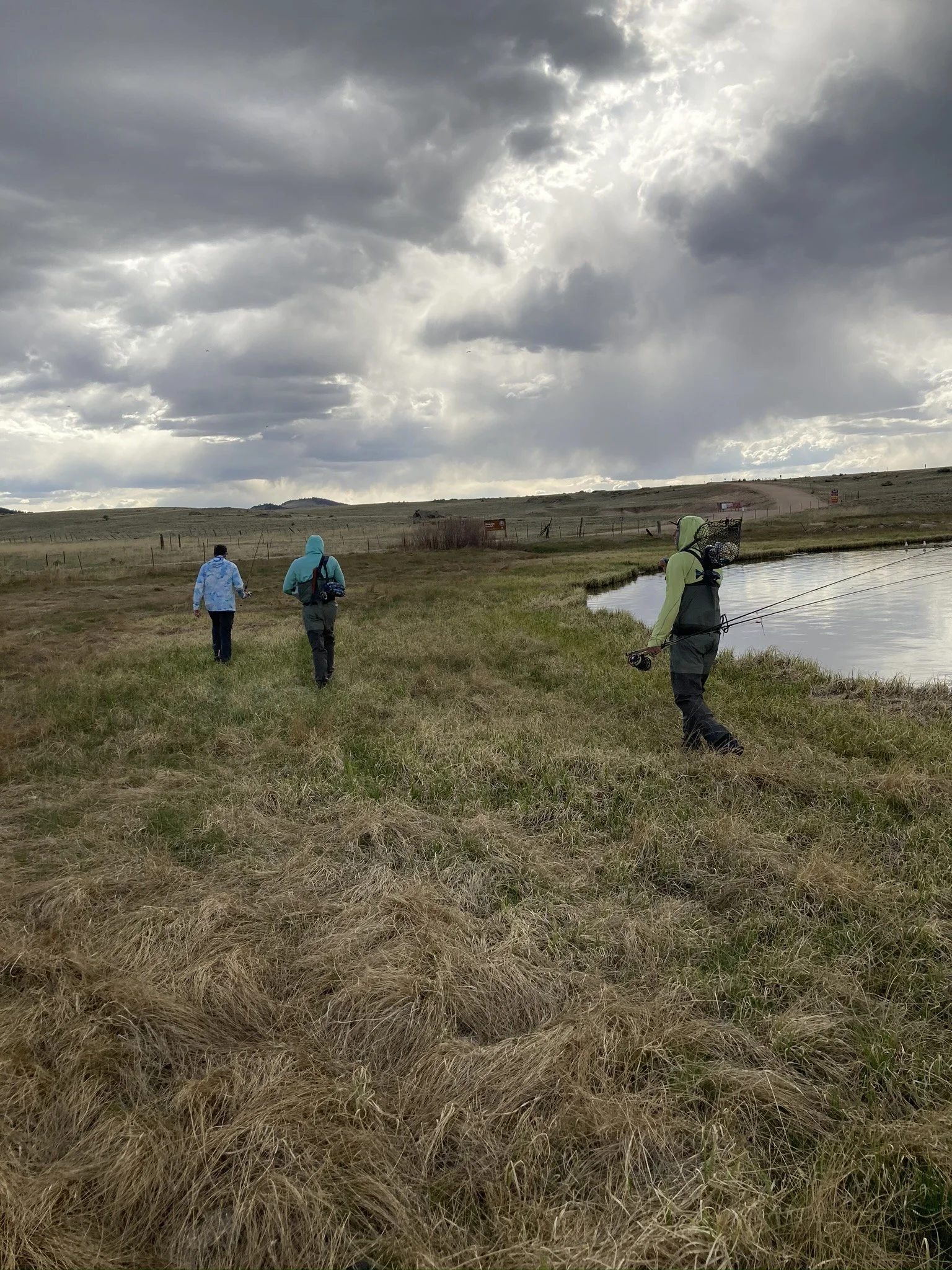 Three people walking along a grassy shoreline by a body of water on a cloudy day, with hills in the background and two fishing rods.