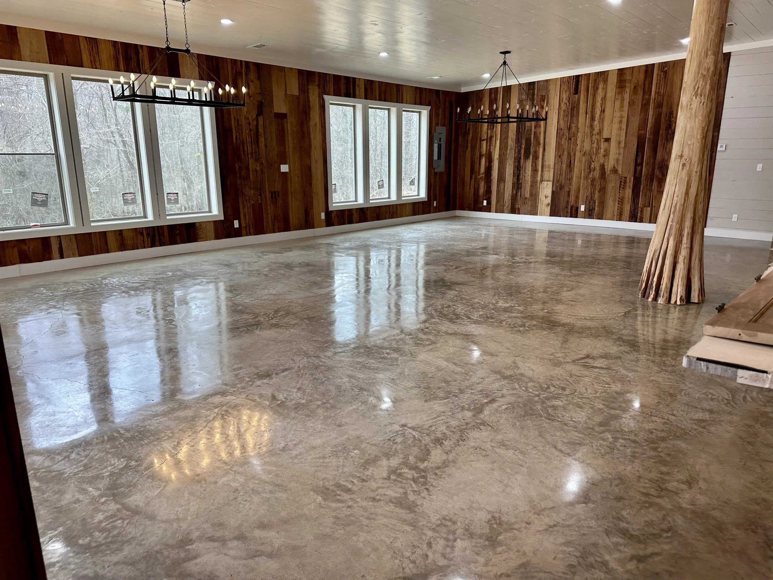 Empty spacious room with polished concrete floor, wood-paneled walls, large windows, and modern chandelier lighting fixtures.