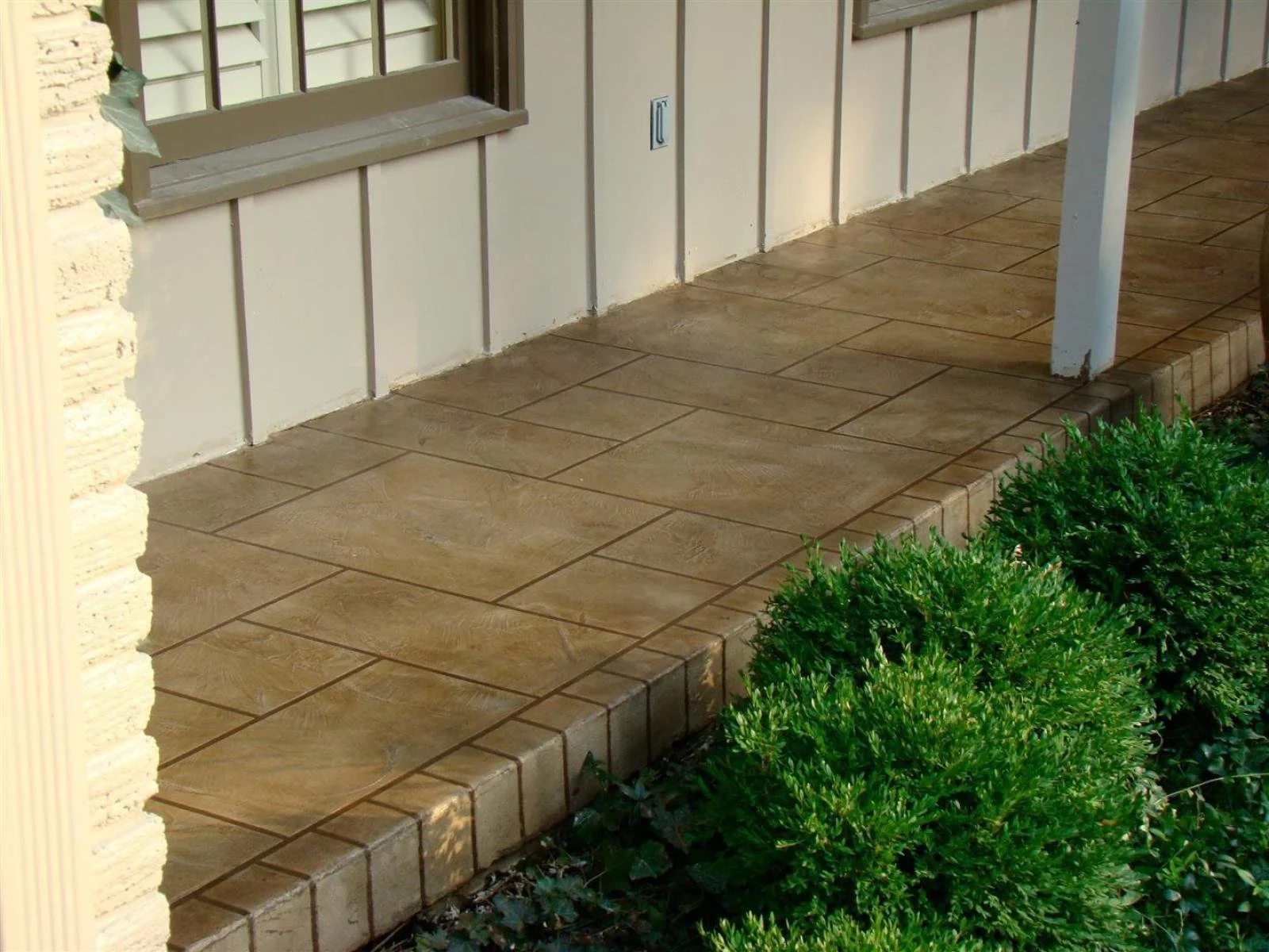 A small front porch with beige tiled flooring, adjacent to a house with beige siding and a window, with green bushes in front.