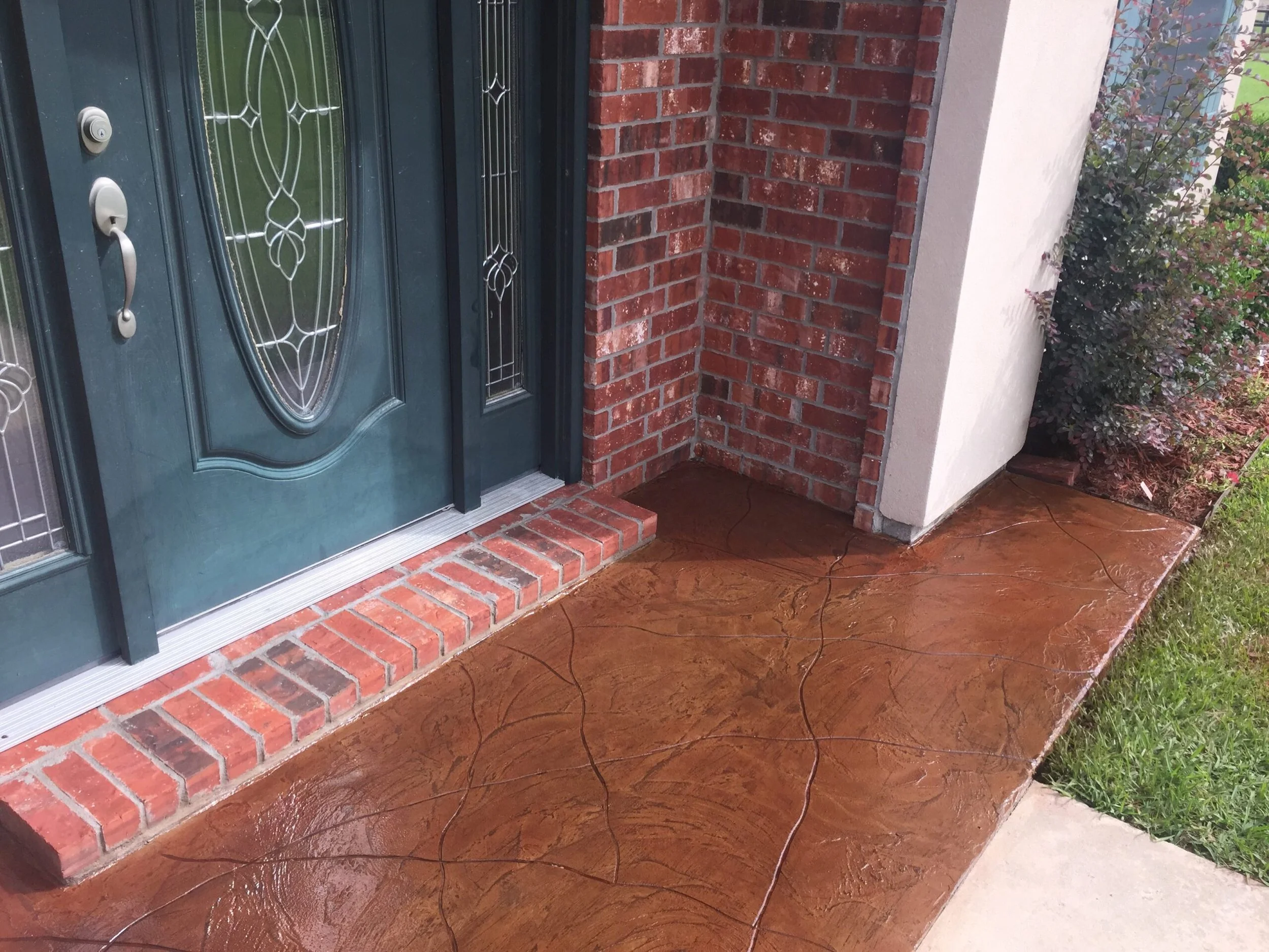 Front porch of a house with a teal front door and stained concrete patio. The door has decorative glass panels and a silver handle. The house has red brick and white siding with some shrubbery nearby.