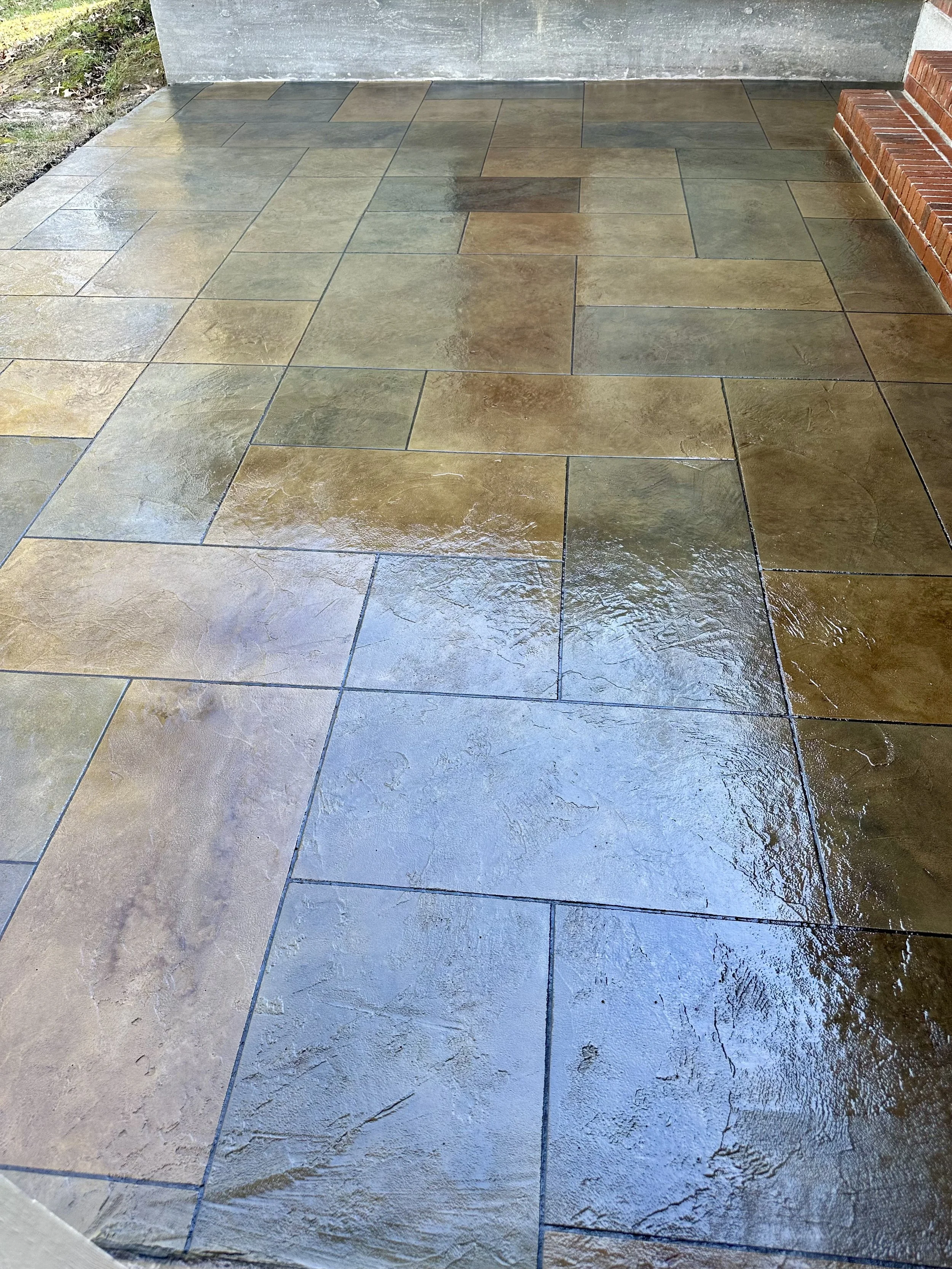 Wet stone patio with rectangular tiles in shades of tan, brown, and gray, adjacent to brick steps and a gray concrete wall.