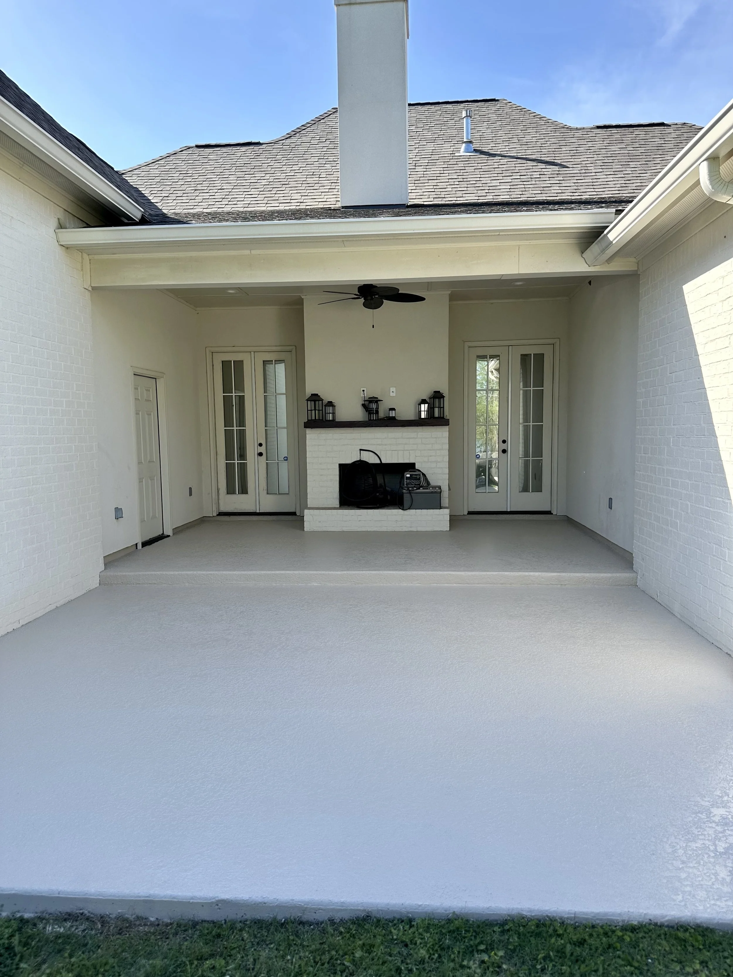 Covered outdoor patio area with white brick walls, beige carpeted floor, ceiling fan, double glass doors, and a built-in white brick fireplace with black lanterns on top.
