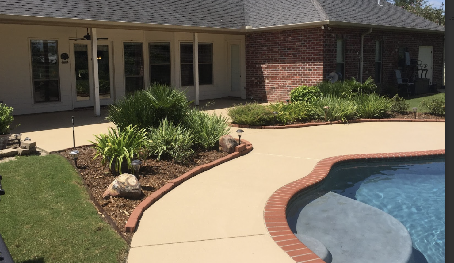 Backyard view showing a concrete patio area with a kidney-shaped pool, landscaped plants and rocks, and a house with brick and siding exterior.
