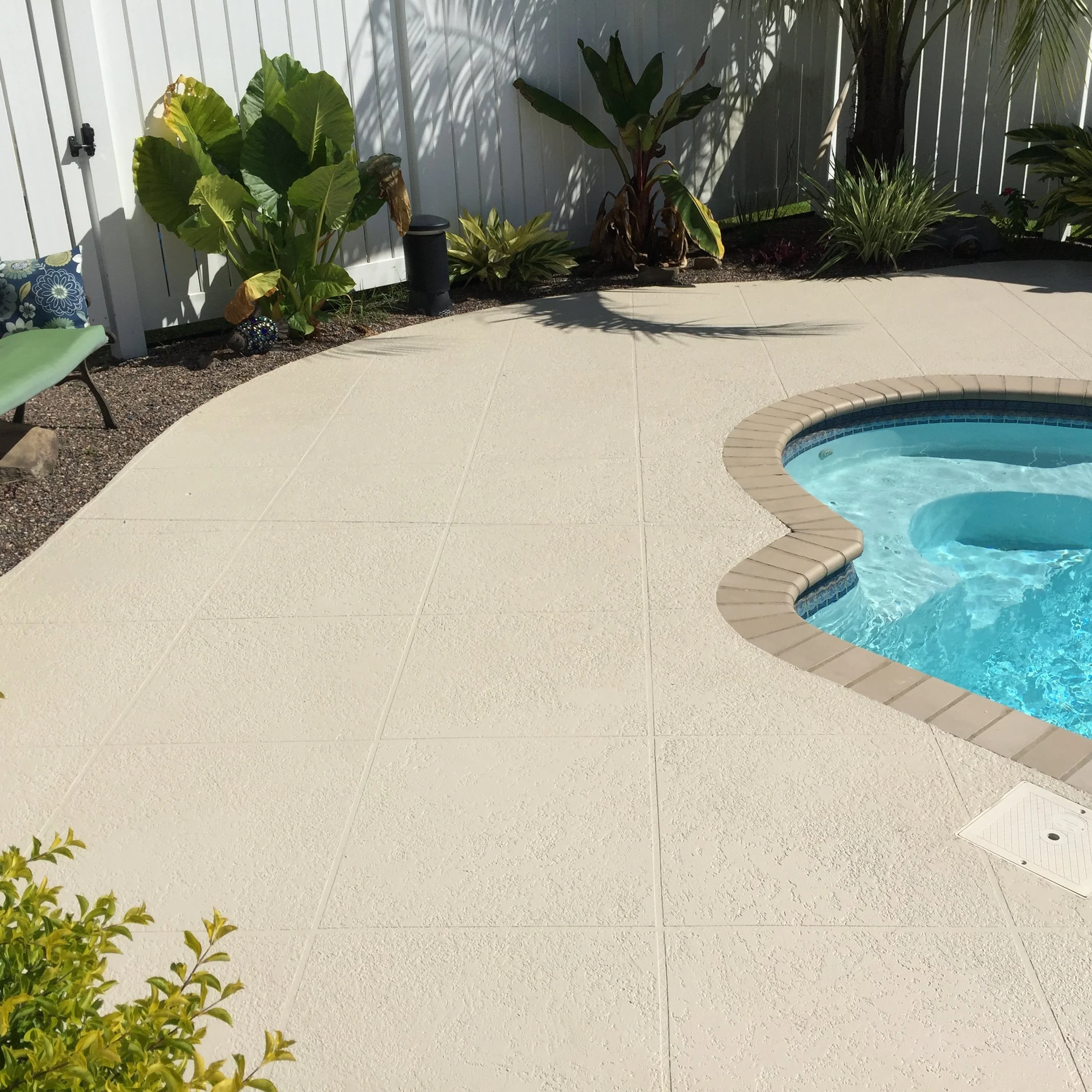 Swimming pool with a curved edge, surrounded by textured concrete patio, with lush green plants and a white fence in the background.