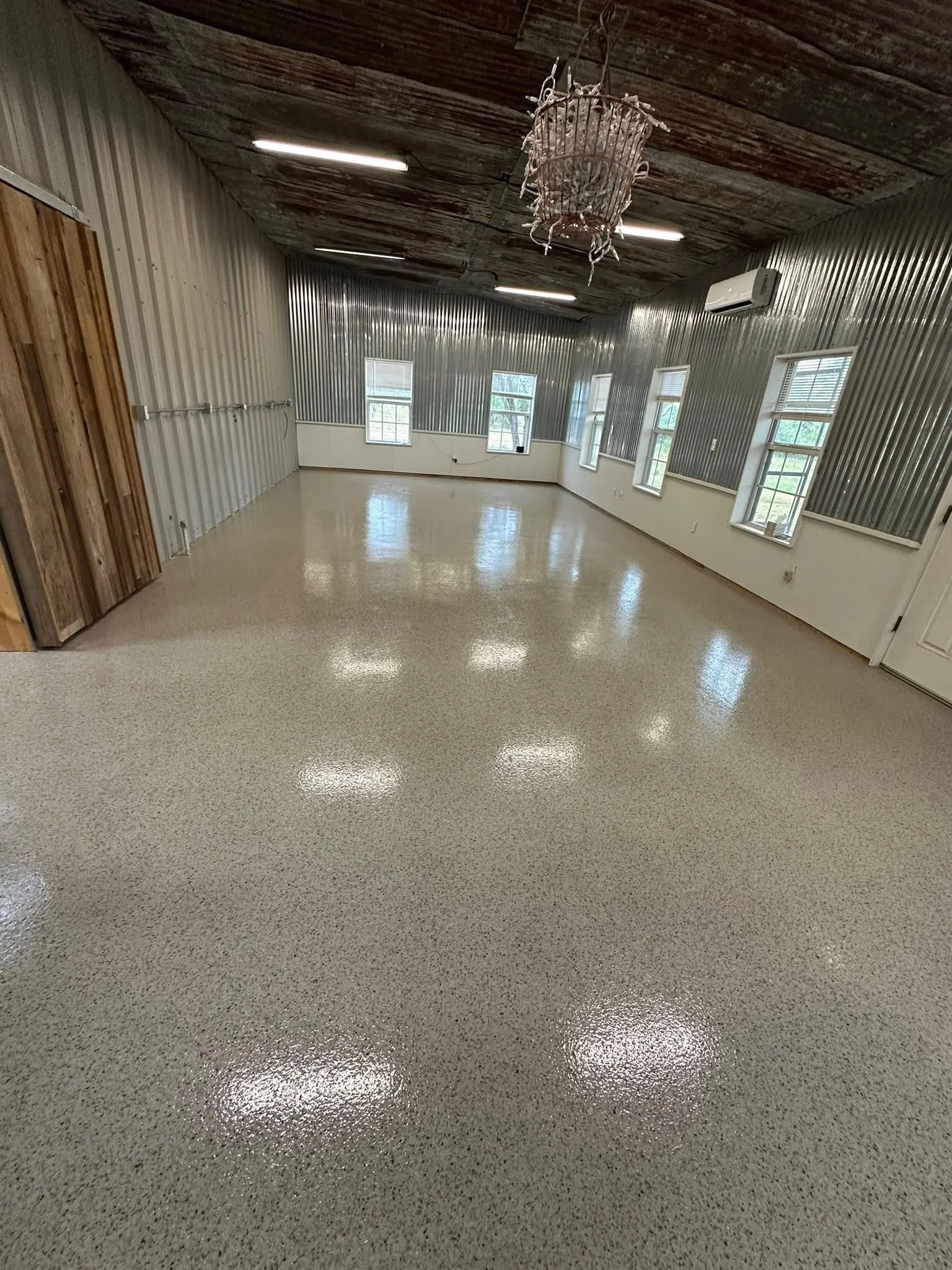 Empty room with polished speckled floor, corrugated metal walls, multiple windows, a decorative chandelier, commercial air conditioning unit, and a wooden door.