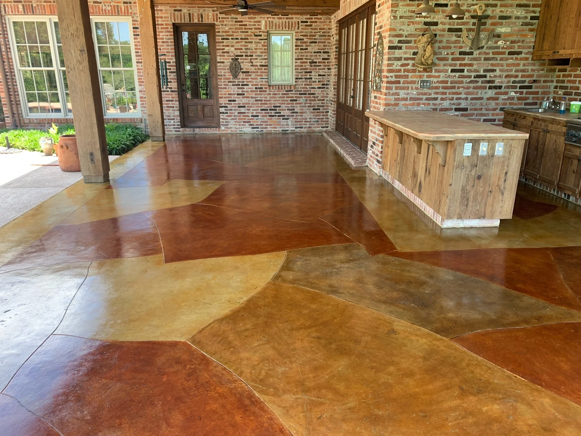 Interior view of a kitchen or porch area with multicolored stained concrete flooring, brick walls, and wooden accents, including a wooden counter and cabinetry.