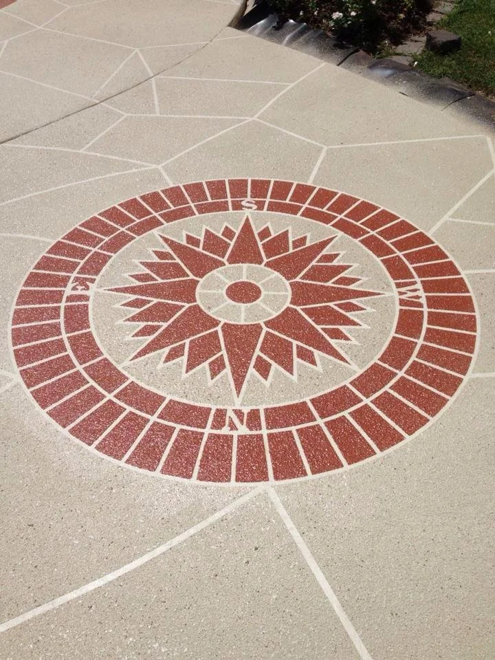 A decorative compass rose design embedded in concrete sidewalk, with cardinal directions marked on a circular pattern in red and white colors.