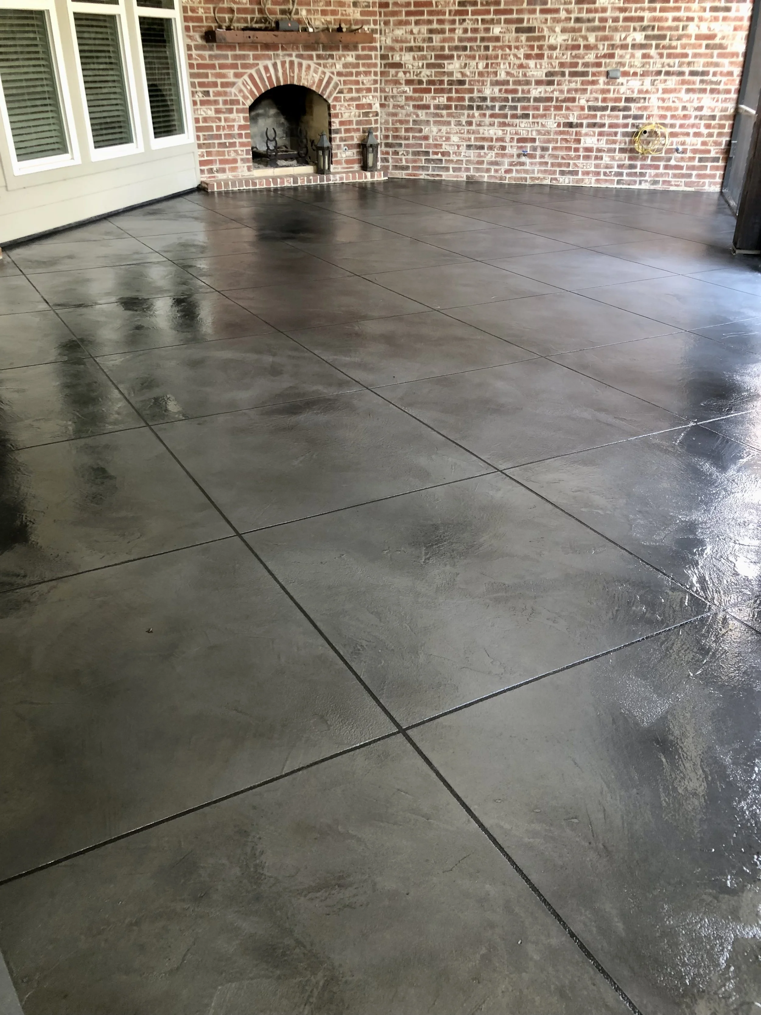 Clean, wet, dark gray concrete patio with visible square tile joints, adjacent to a brick wall with a fireplace, and a window with white blinds.
