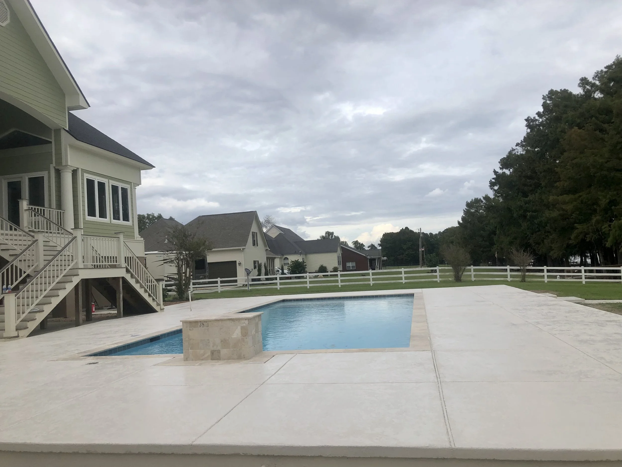 Residential backyard with a rectangular swimming pool surrounded by a concrete deck, adjacent to a two-story house with a covered porch and stairs. Other houses and a white fence are visible in the background under a cloudy sky.
