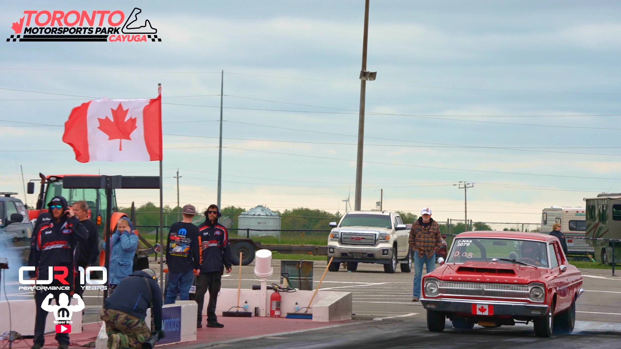 A drag racing event at Toronto Motorsports Park Cayuga with a red vintage car, likely a Plymouth, at the starting line, a large Canadian flag waving, and several people in racing gear and casual clothing standing nearby. Overcast sky and racing facilities in the background.