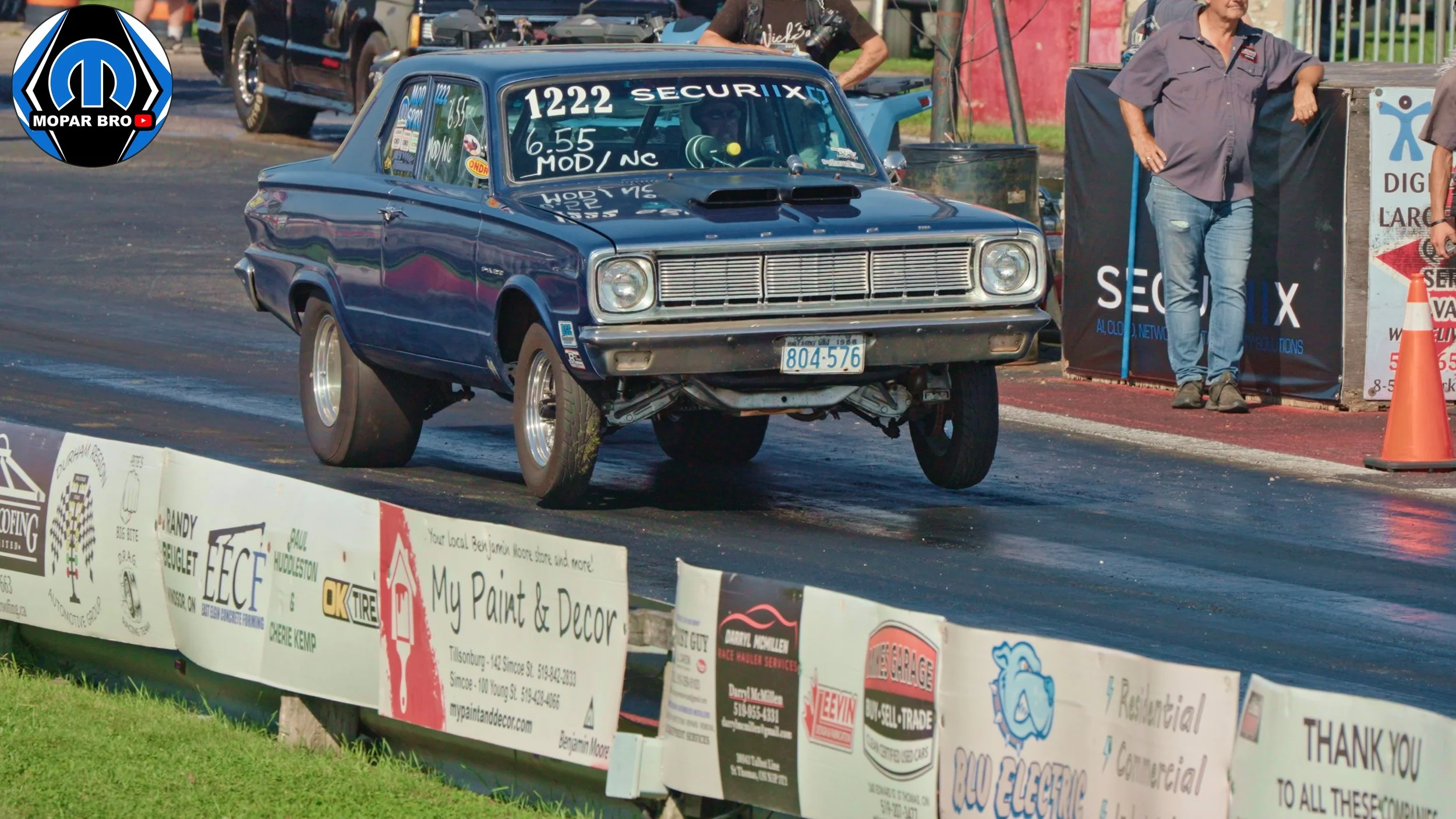 A vintage blue Dodge dart drag racing car with raised front suspension, racing on a drag strip with racing stickers and sponsor logos. Drag racing at the st thomas dragway in st thomas ontario