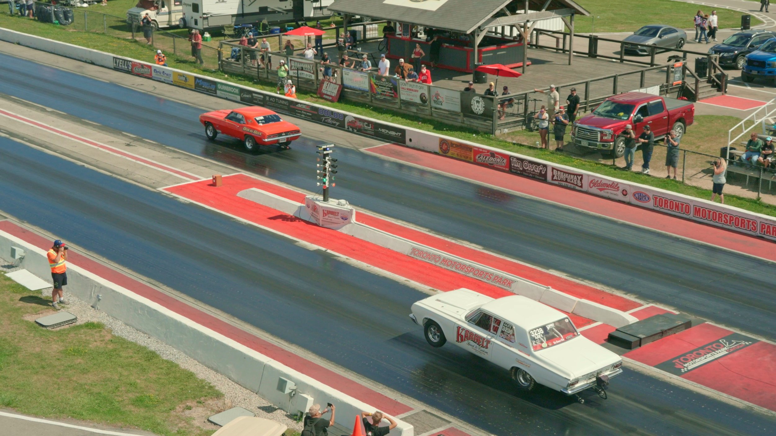 A drag racing event at Toronto Motorsports Park featuring two classic muscle cars, one red and one white, on the race strip with spectators watching from the stands and along the fence. plymouth belvedere drag race