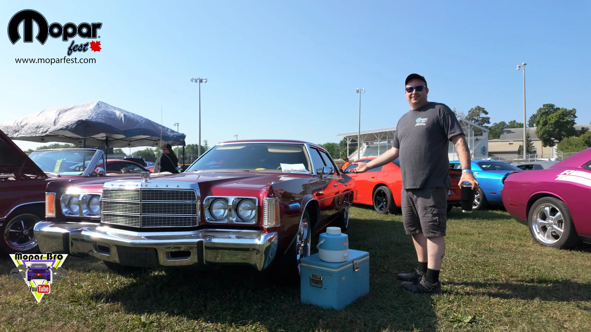 A man standing next to a classic maroon car at a Mopar car show, with other colorful vintage cars and tents in the background on a sunny day at moparfest beside a chrysler newport