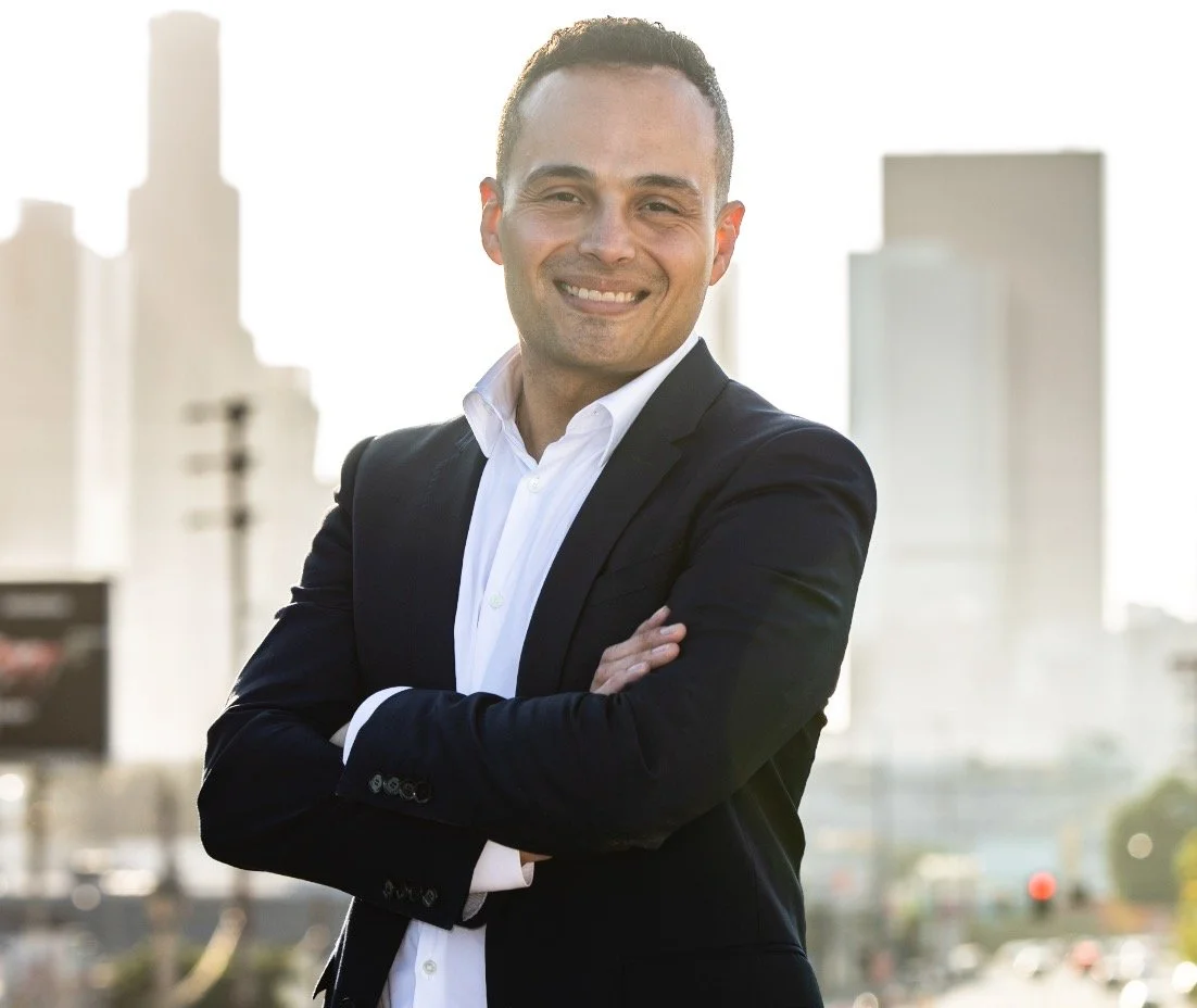 A man in a black suit and white shirt standing outdoors with arms crossed, smiling, with city buildings in the background.