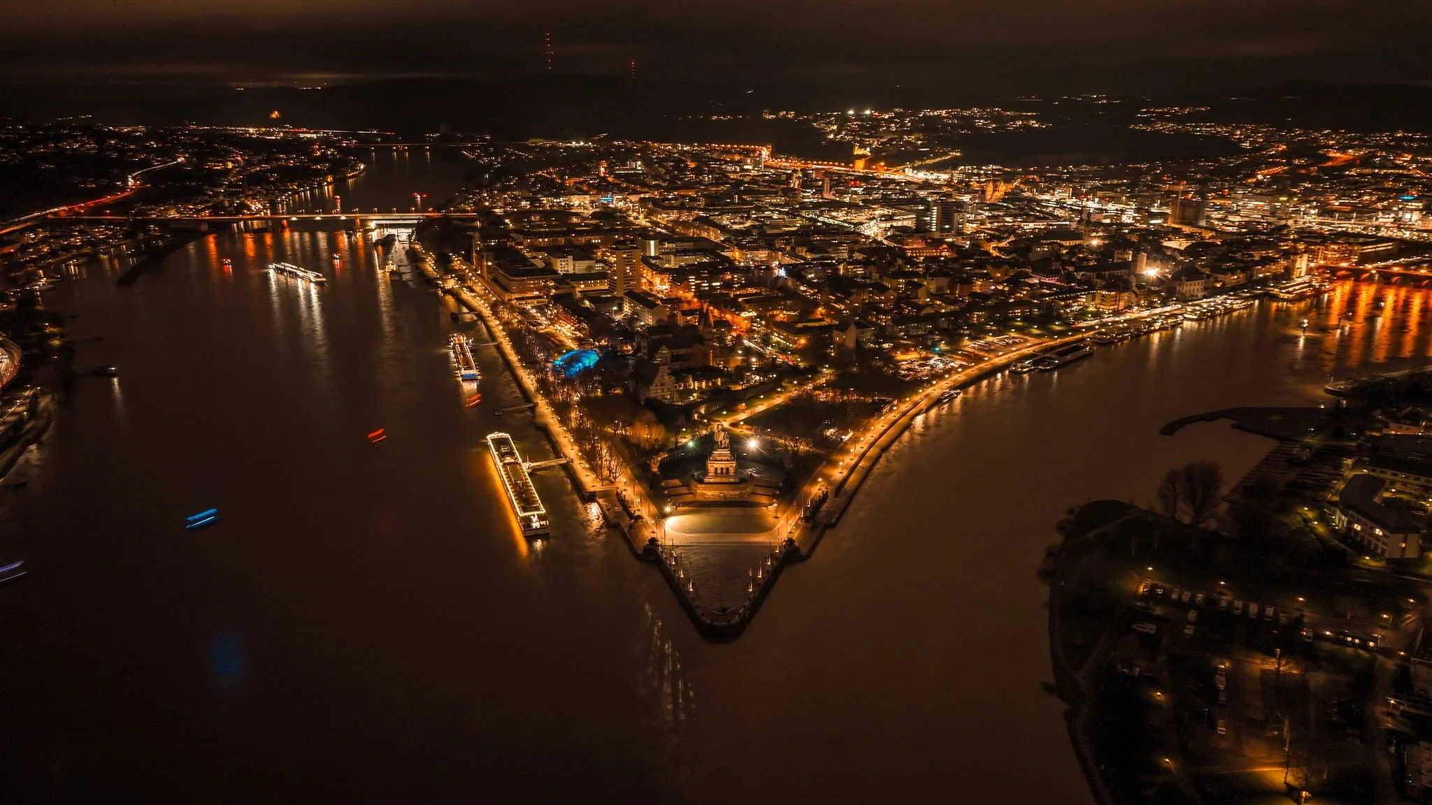 An aerial night view of a cityscape featuring a river with boats, illuminated streets, and buildings, with a prominent triangular park or landmark at the point of the river.