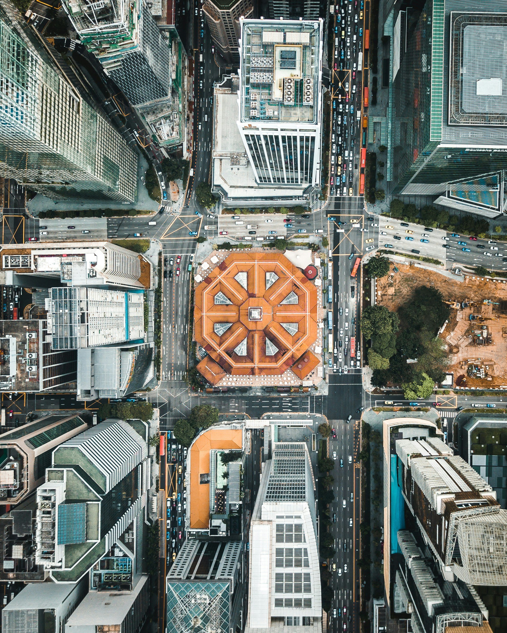 An aerial view of a city intersection with tall buildings, roads, traffic, a large orange-roofed building at the center, and some trees and construction site on the right.