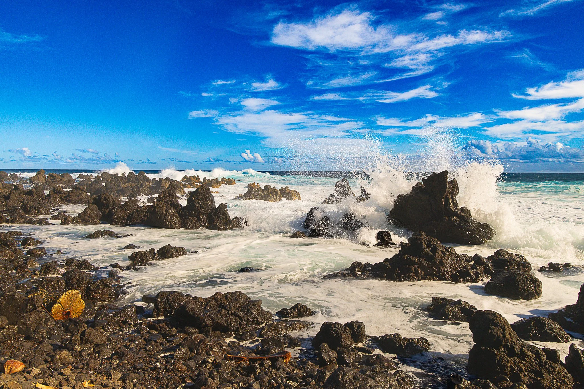 Oceanside view with rocks and crashing waves under a blue sky with scattered clouds.