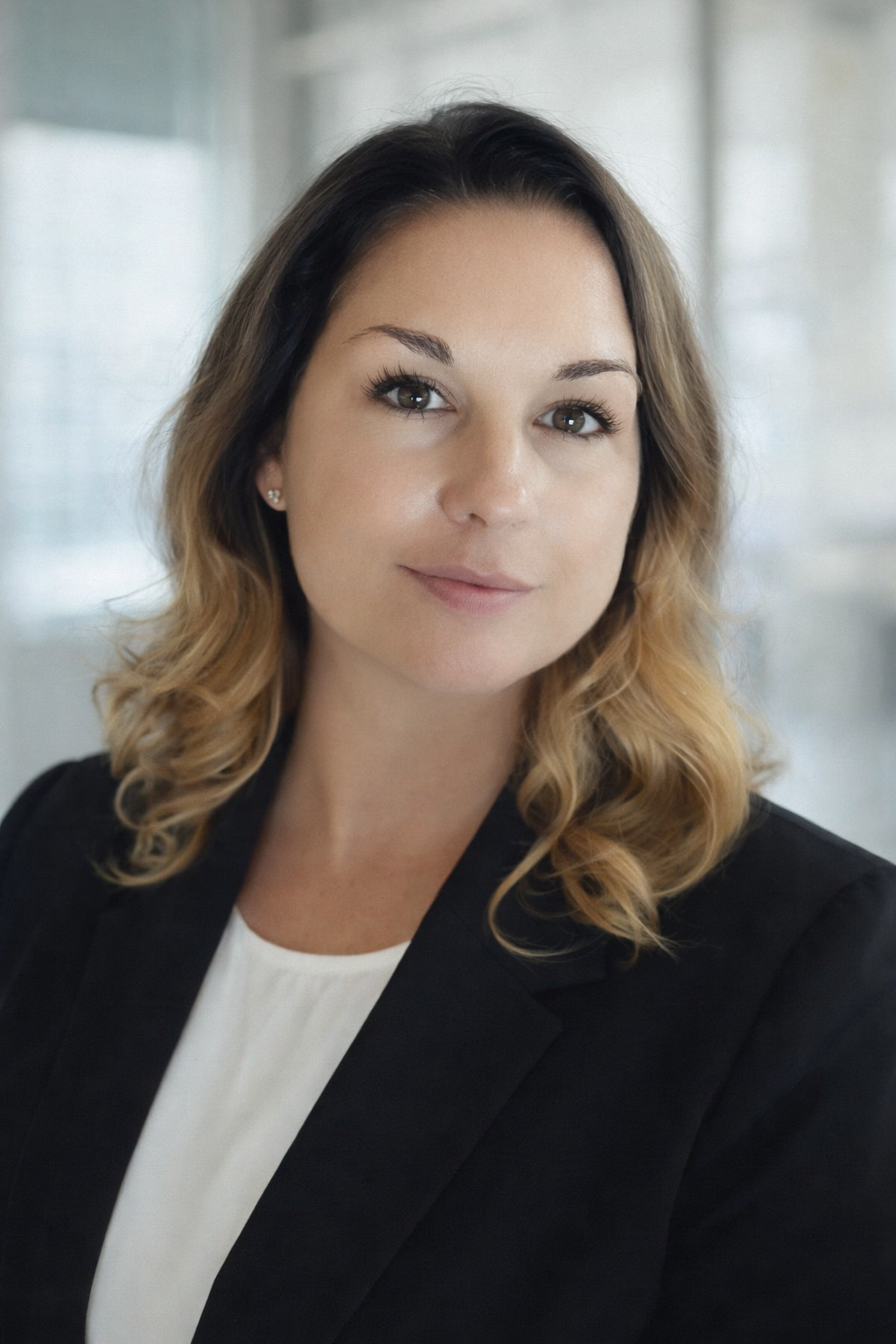 A professional woman with shoulder-length wavy hair, wearing a black blazer and white top, smiling softly in a modern office environment.