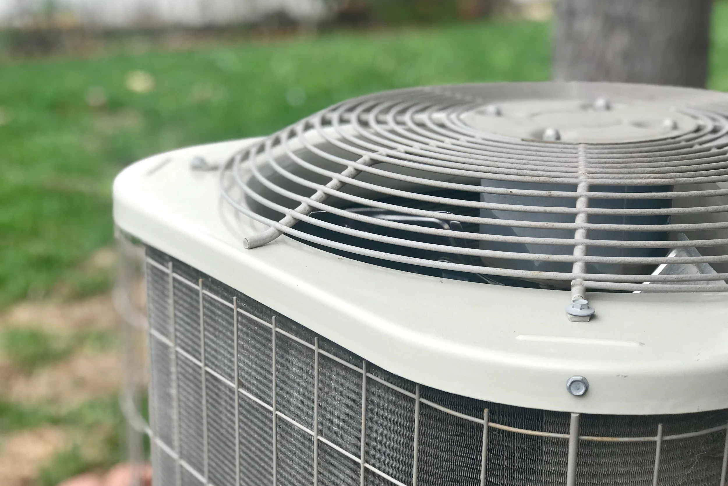 Close-up of an outdoor air conditioning unit with a ventilation fan and metal grille in a grassy yard.