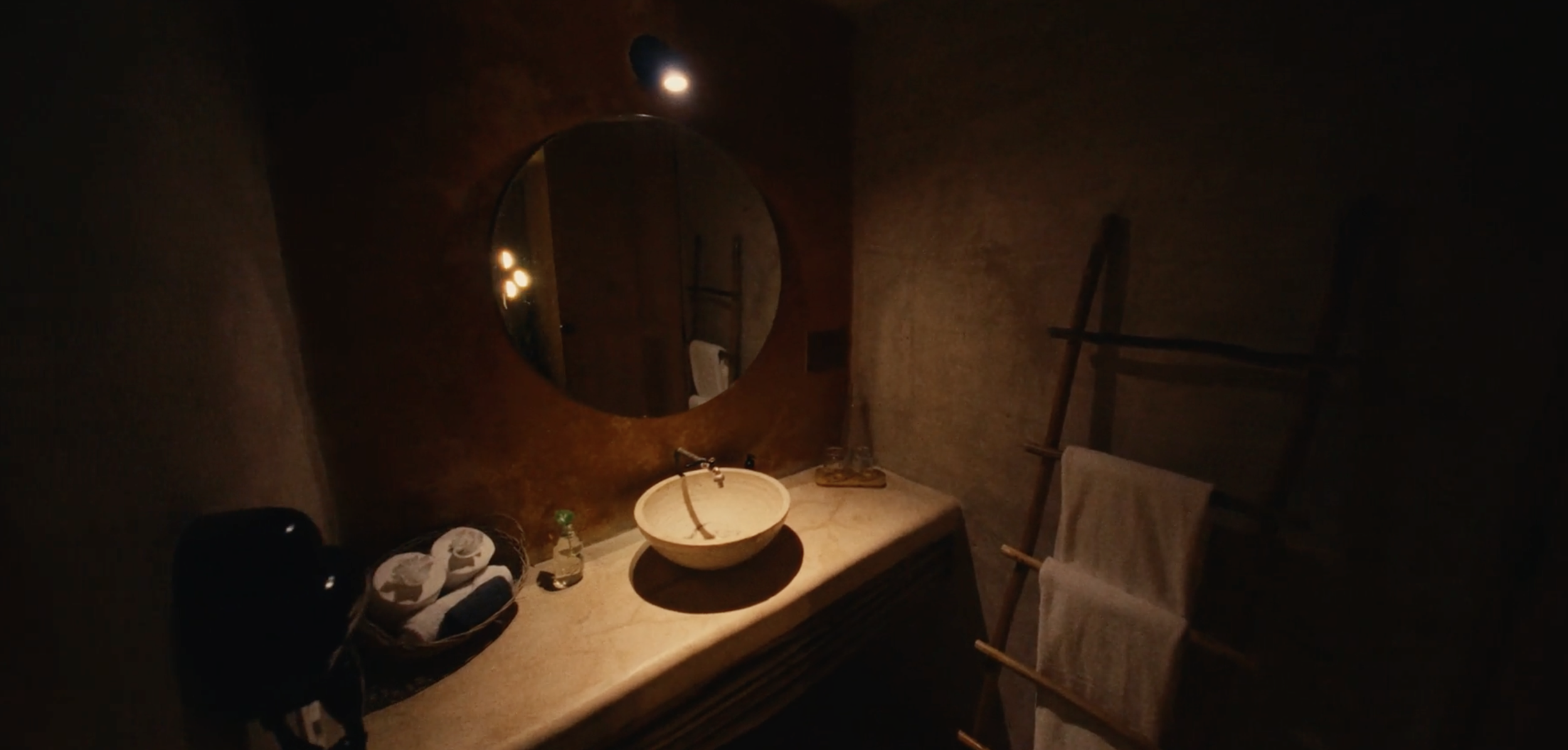 Vista de un baño con lavabo de piedra, espejo redondo, cestas con toallas, planta pequeña y escalera rack con toallas. La iluminación es tenue y cálida.