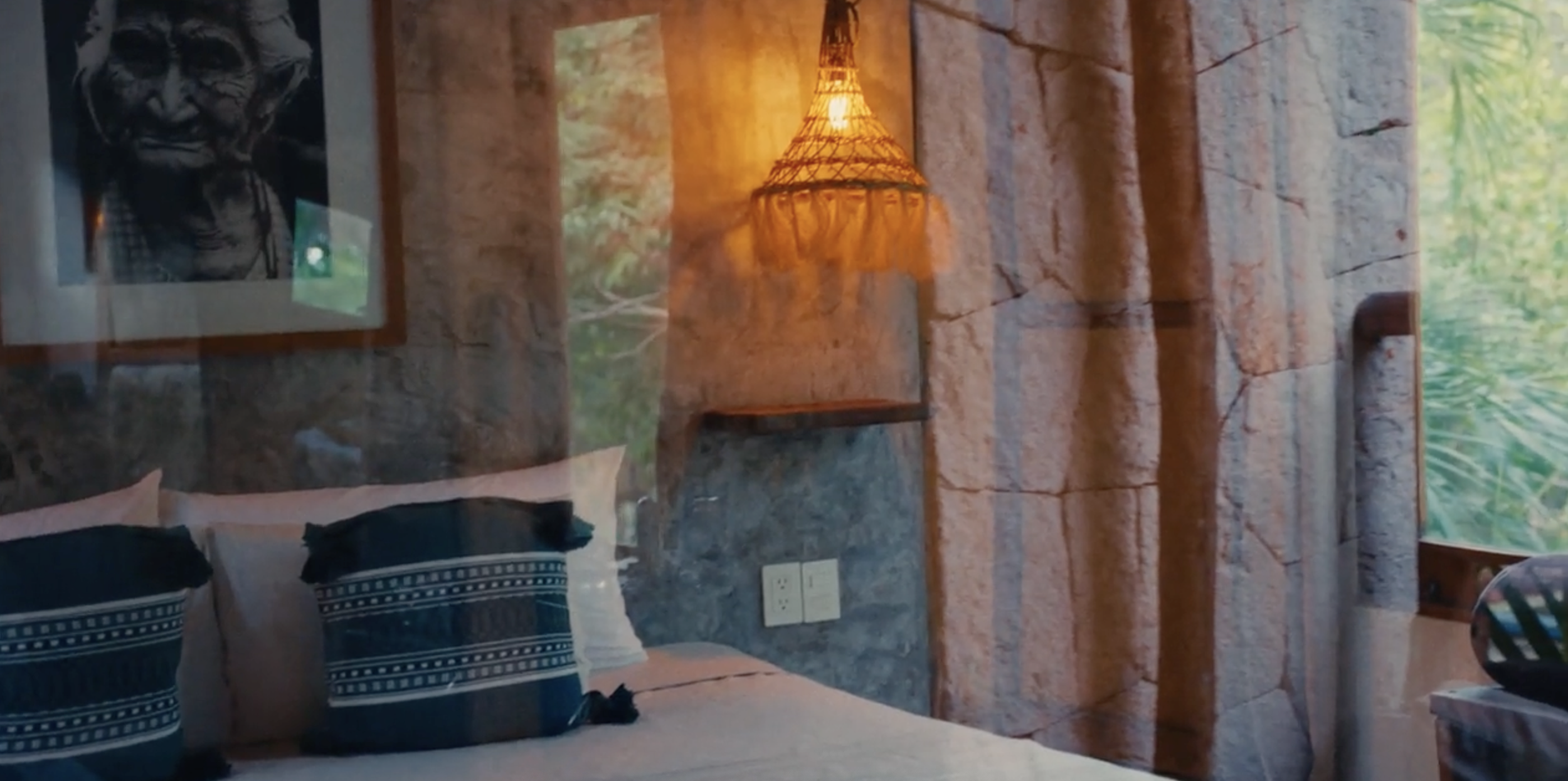 Habitación con paredes de piedra, dos camas con almohadas negras, un cuadro con retrato de un tigre, lámpara de mimbre, ventana con vista a vegetación verde.