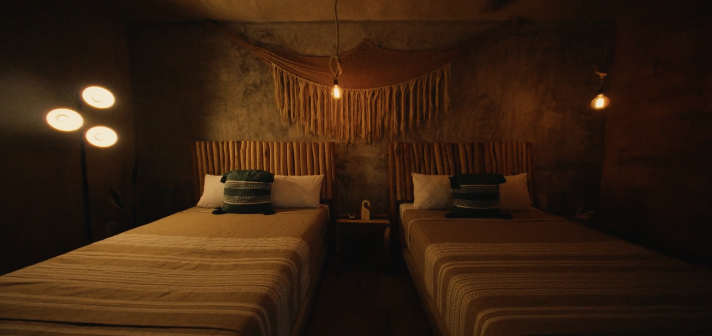 Dos camas en un dormitorio con paredes de piedra, cabezales de madera y decoración de tela en tonos cálidos, iluminación tenue y lámparas en el techo.