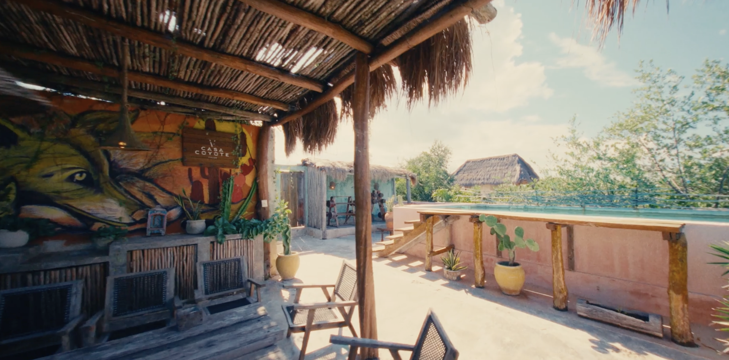 Área exterior con muebles de madera, plantas en macetas y un mural colorido; estructura de techo con paredes de madera, con espacio para descanso y relajación.