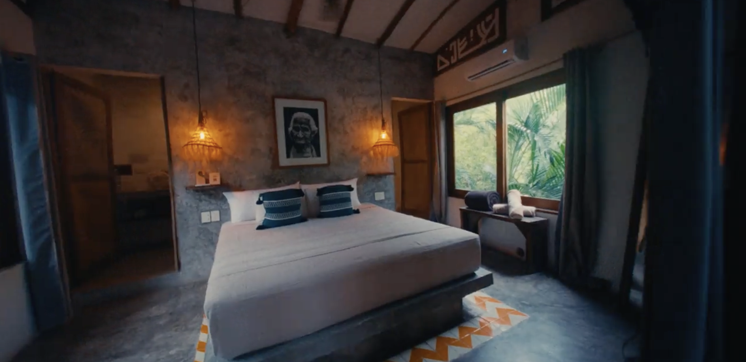 Habitación con cama, paredes de piedra, ventana grande con vista a plantas verdes, lámparas colgantes de madera, foto de perfil en la pared, cortinas y aire acondicionado.