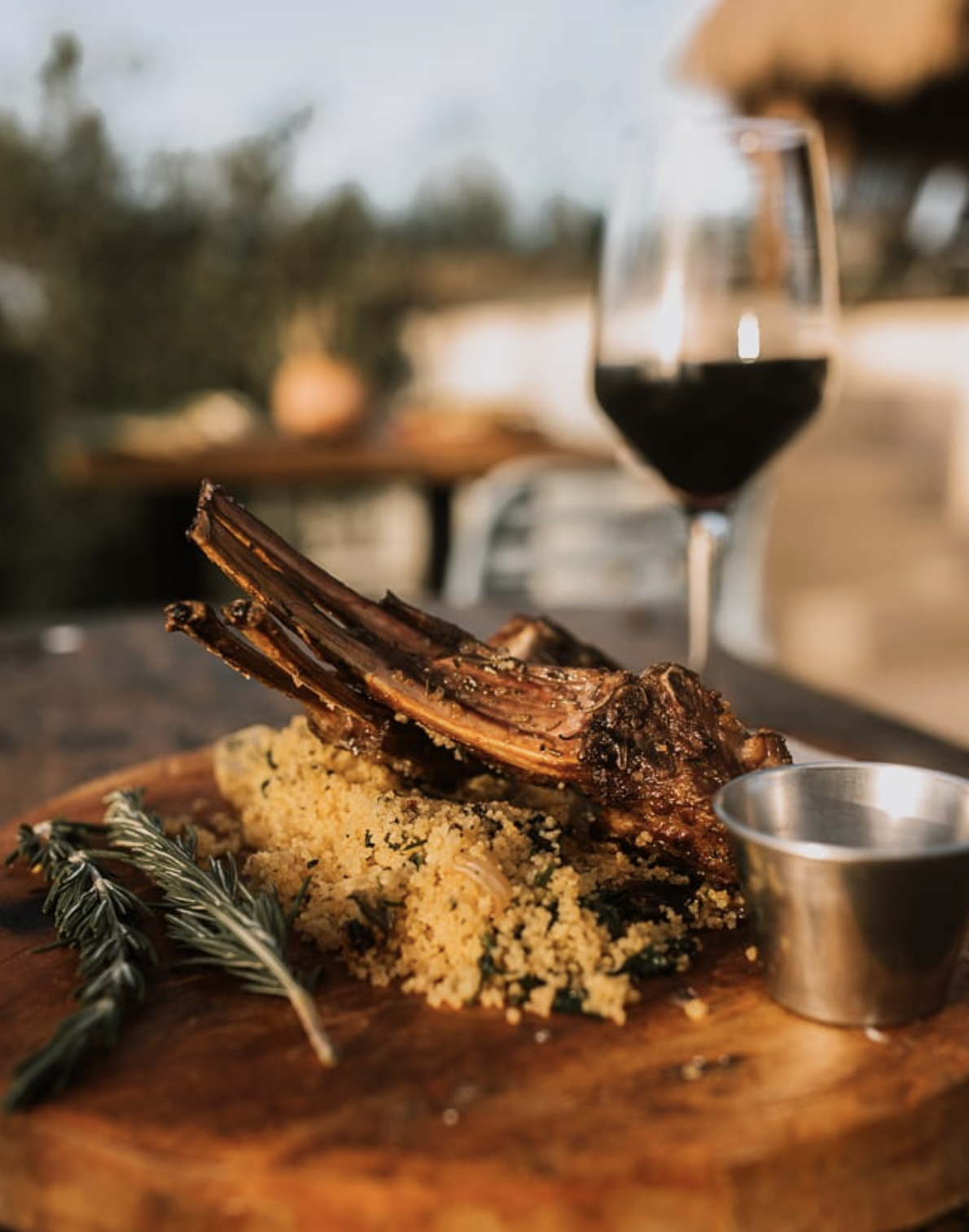 Cordero asado en plato de madera con un lado de cuscús y ramitas de romero. Vino tinto en copa al fondo.
