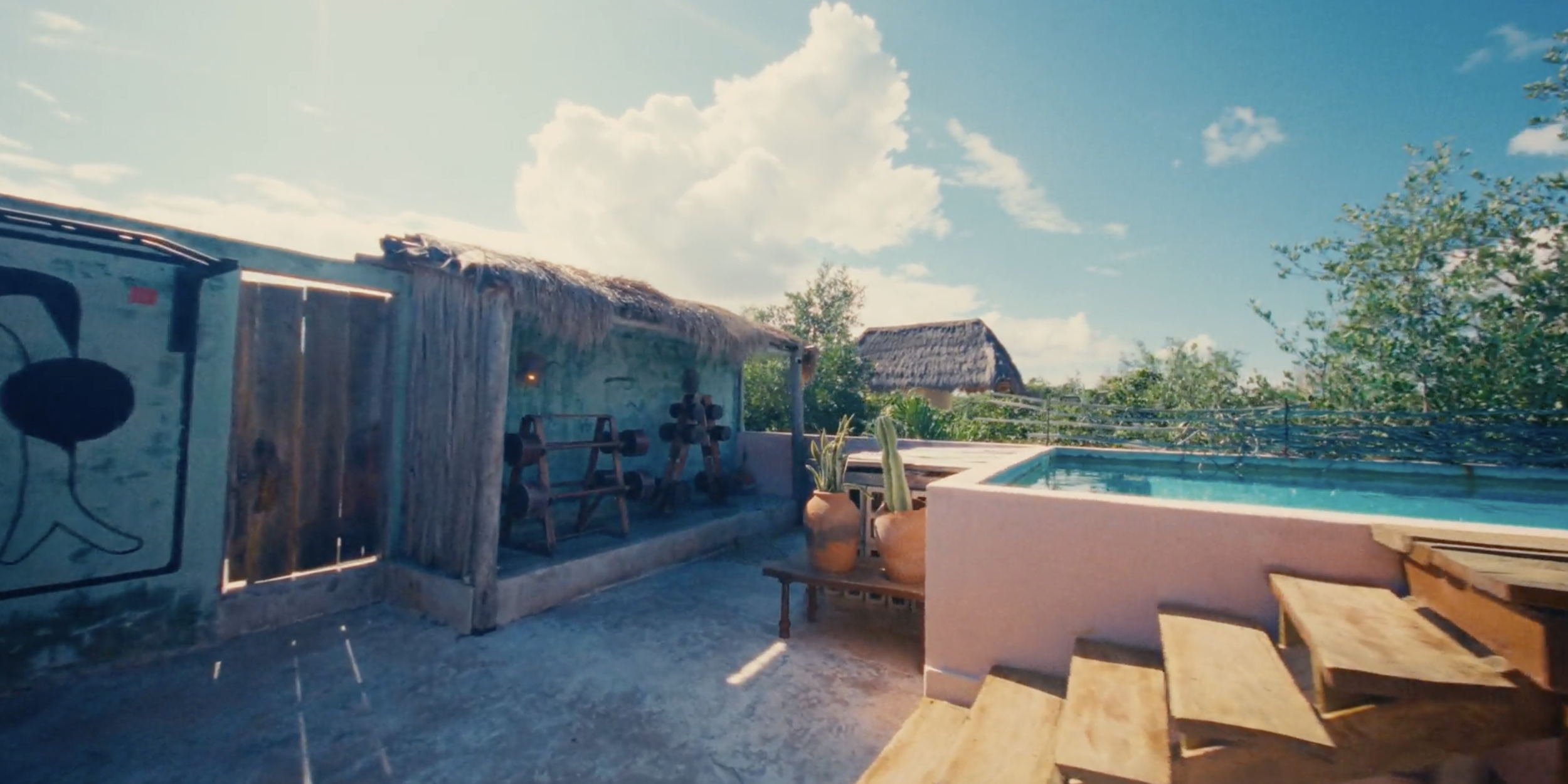 Piscina al aire libre con silla y plantas en jarrón, cercado de madera y casas tradicionales en el fondo bajo cielo despejado.