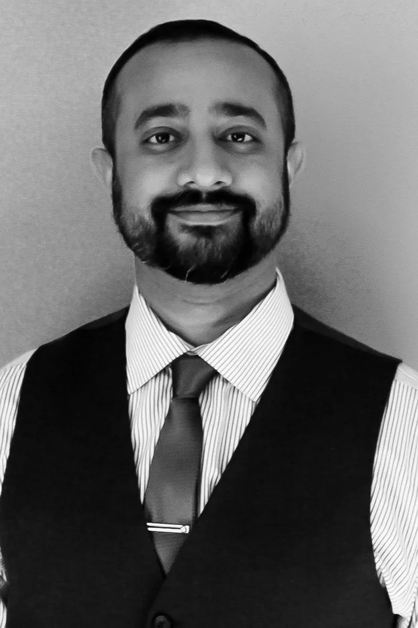 Portrait of a man with a beard and mustache wearing a suit vest, striped shirt, and tie, smiling in front of a plain background.