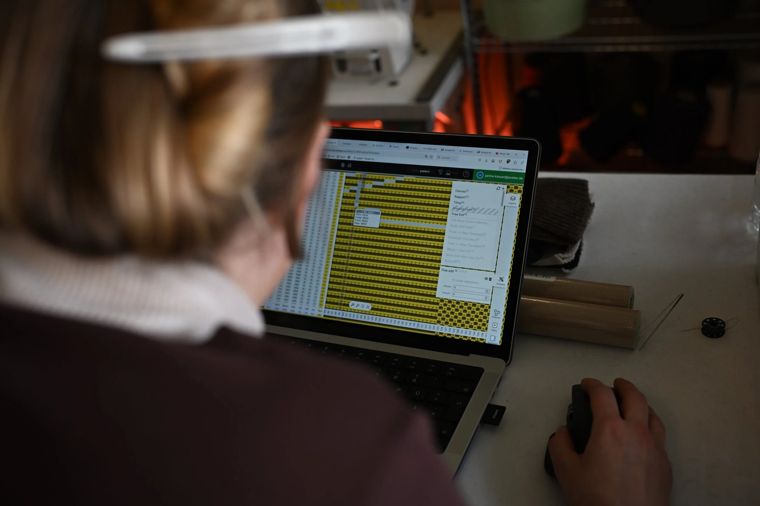 Eine Person arbeitet an einem Laptop, der eine Tabelle mit gelben Zeilen anzeigt, vermutlich in einem Büro oder einer Werkstatt, mit Werkzeugen und anderen Gegenständen auf dem Tisch.