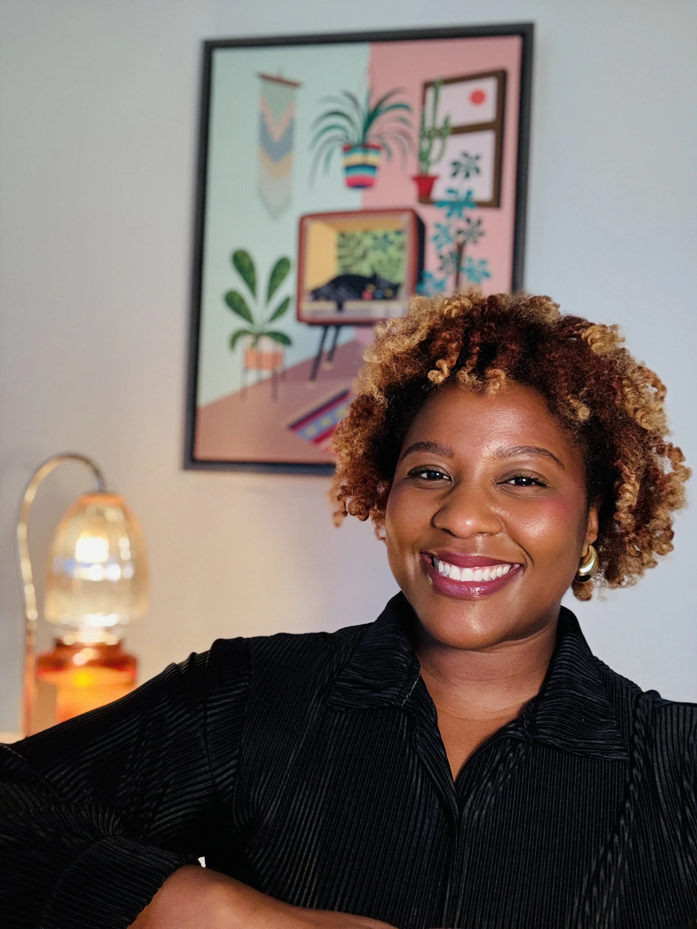 A woman with curly, reddish-brown hair taking a selfie indoors, smiling with white teeth, wearing a black collared top, with a colorful framed artwork and a lit lamp in the background.