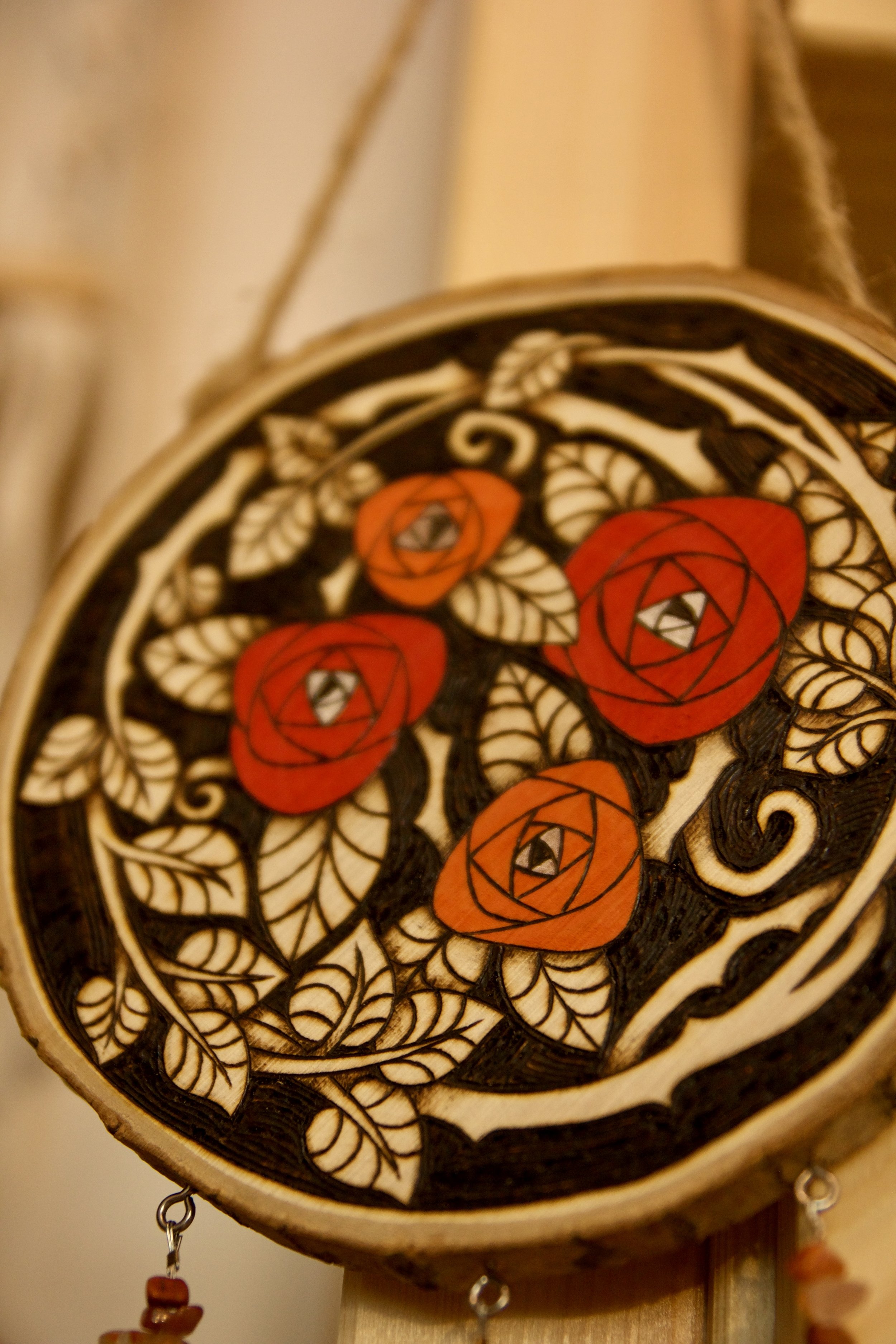 Détail de texture du bois d'une amulette murale ronde en bois pyrogravé et peint avec des roses rouges, ornée de pendants en pierre et d'une rose en céramique.