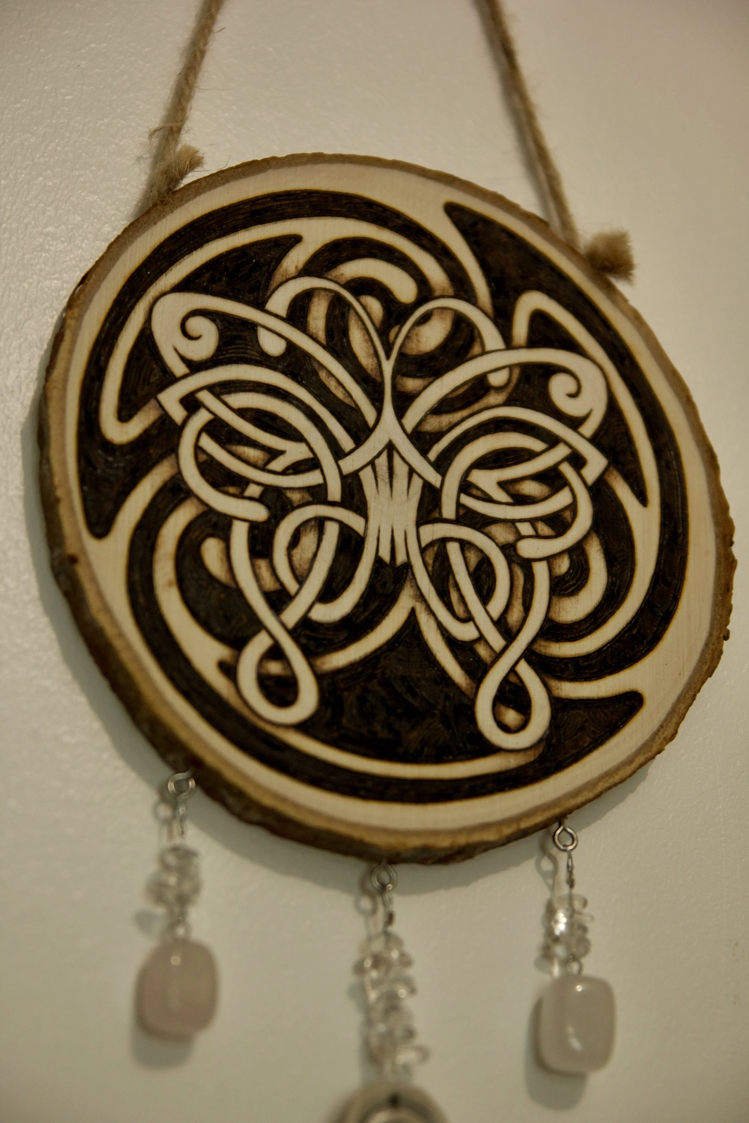 Zoom sur une amulette murale artisanale en bois pyrogravé représentant un papillon mêlé à un triskèle celte, ornée de céramique et de minéraux.