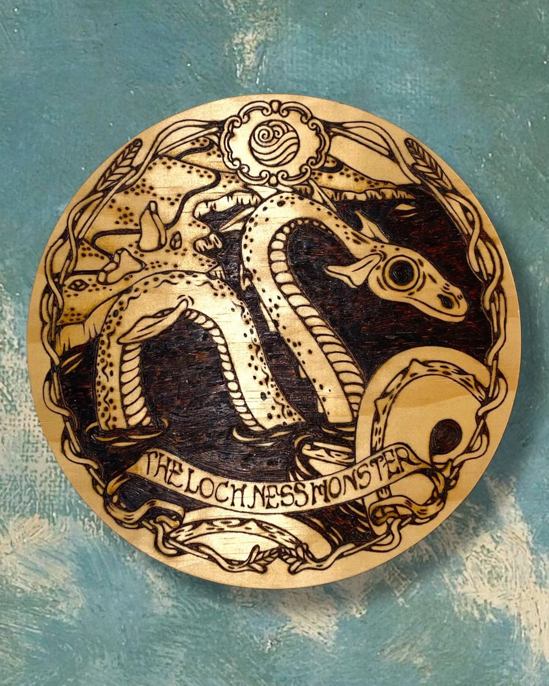 Pyrogravure artisanale ronde sur bois représentant la silhouette mystique du monstre du Loch Ness sous les eaux sombres d'Écosse sur un fond de nuages.
