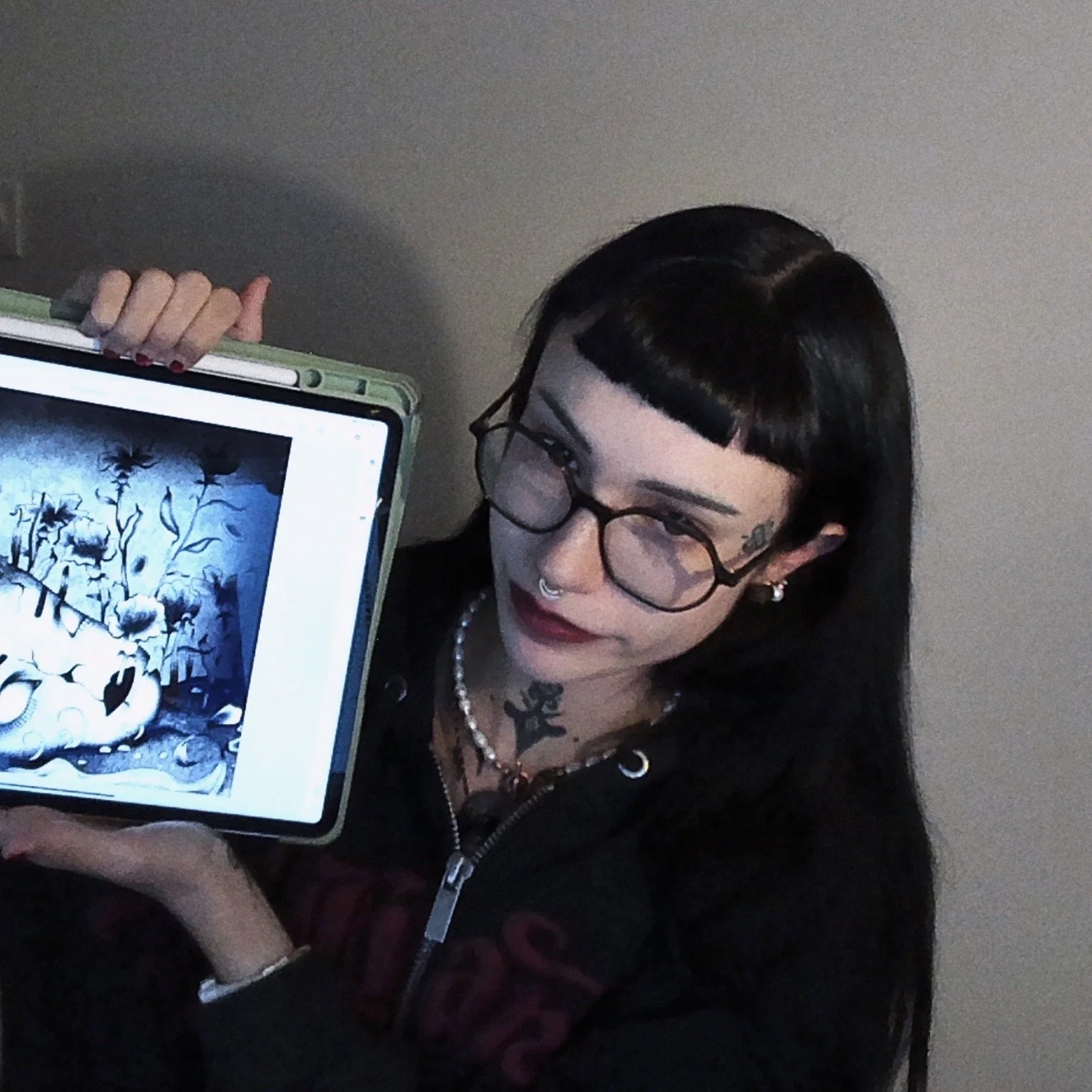Une femme avec des tatouages, des lunettes rondes, un piercing au nez, tient une tablette affichant une illustration d'une scène avec des fleurs.