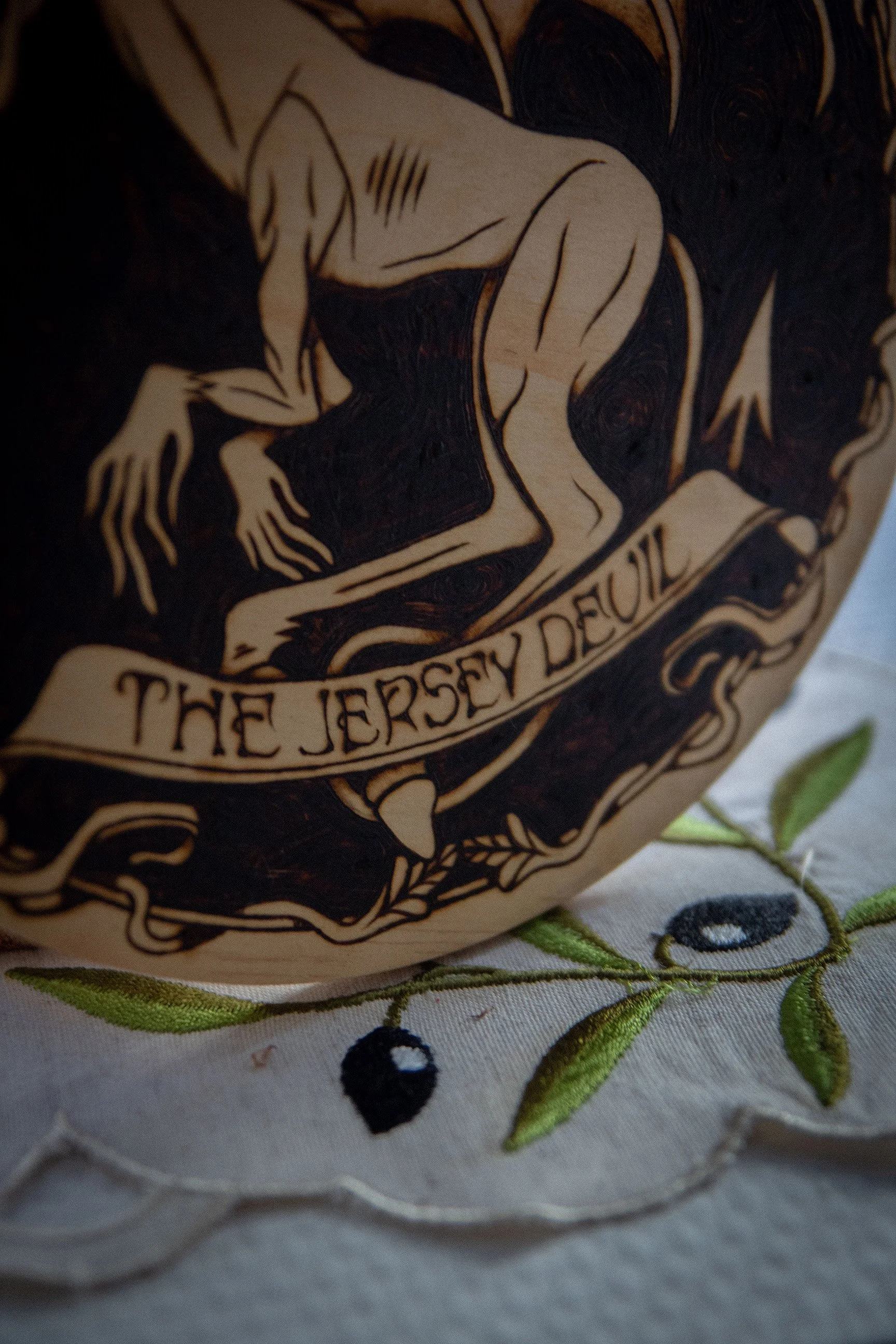 Détail de typographie d'une pyrogravure artisanale ronde de 20x20 cm sur bois représentant la silhouette du Diable de Jersey, créature ailée mythique du folklore américain.