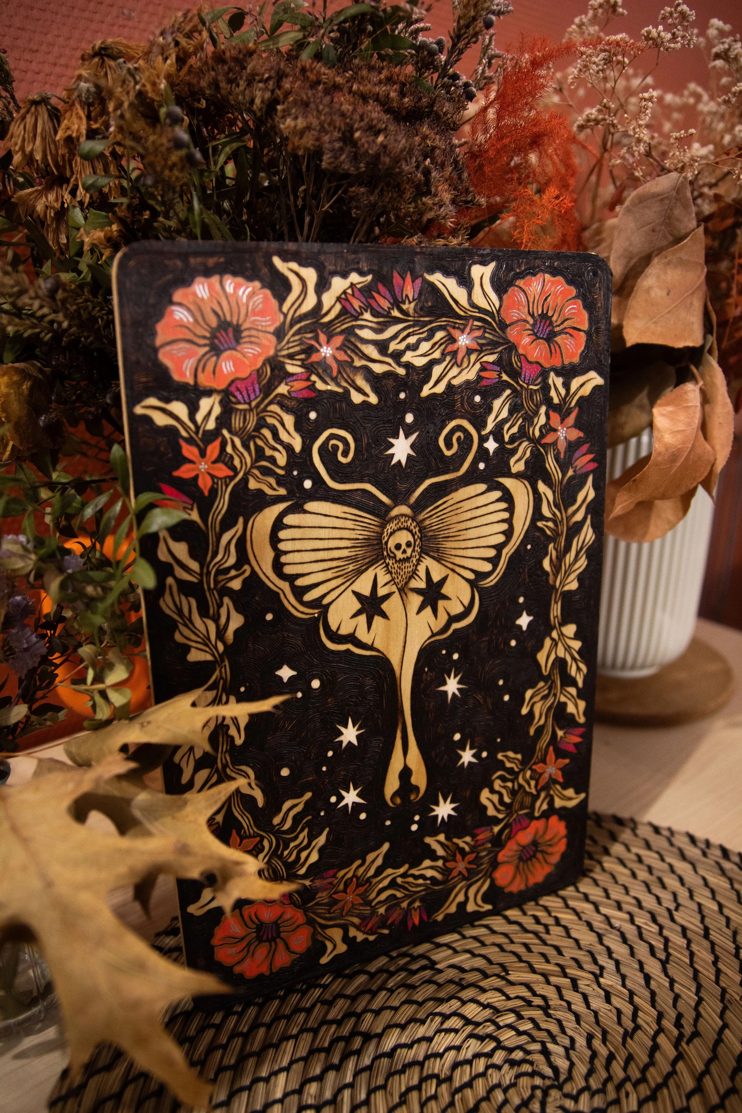 Plaque en bois artisanale pyrogravée d'un papillon de nuit mystique entouré de fleurs délicates sublimées par des touches de peinture colorée.