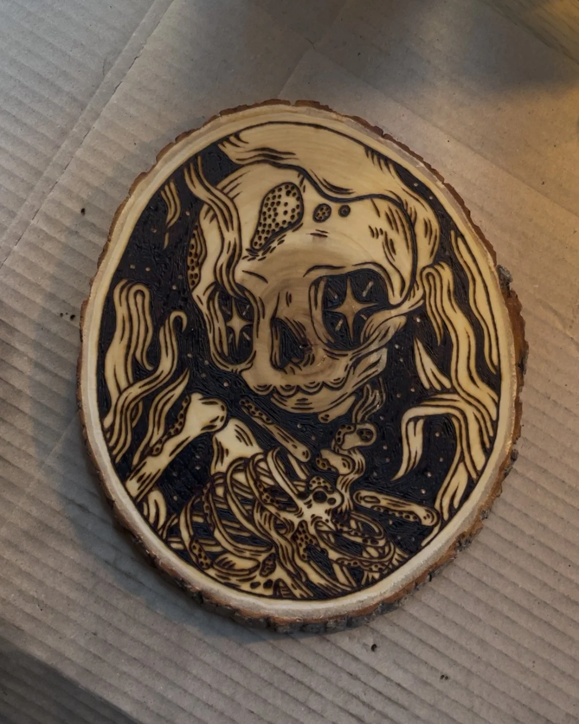 Pyrogravure artisanale sur bois brut ovale représentant un squelette Memento Mori au style d'un artefact ancien sur un bureau d'artiste.
