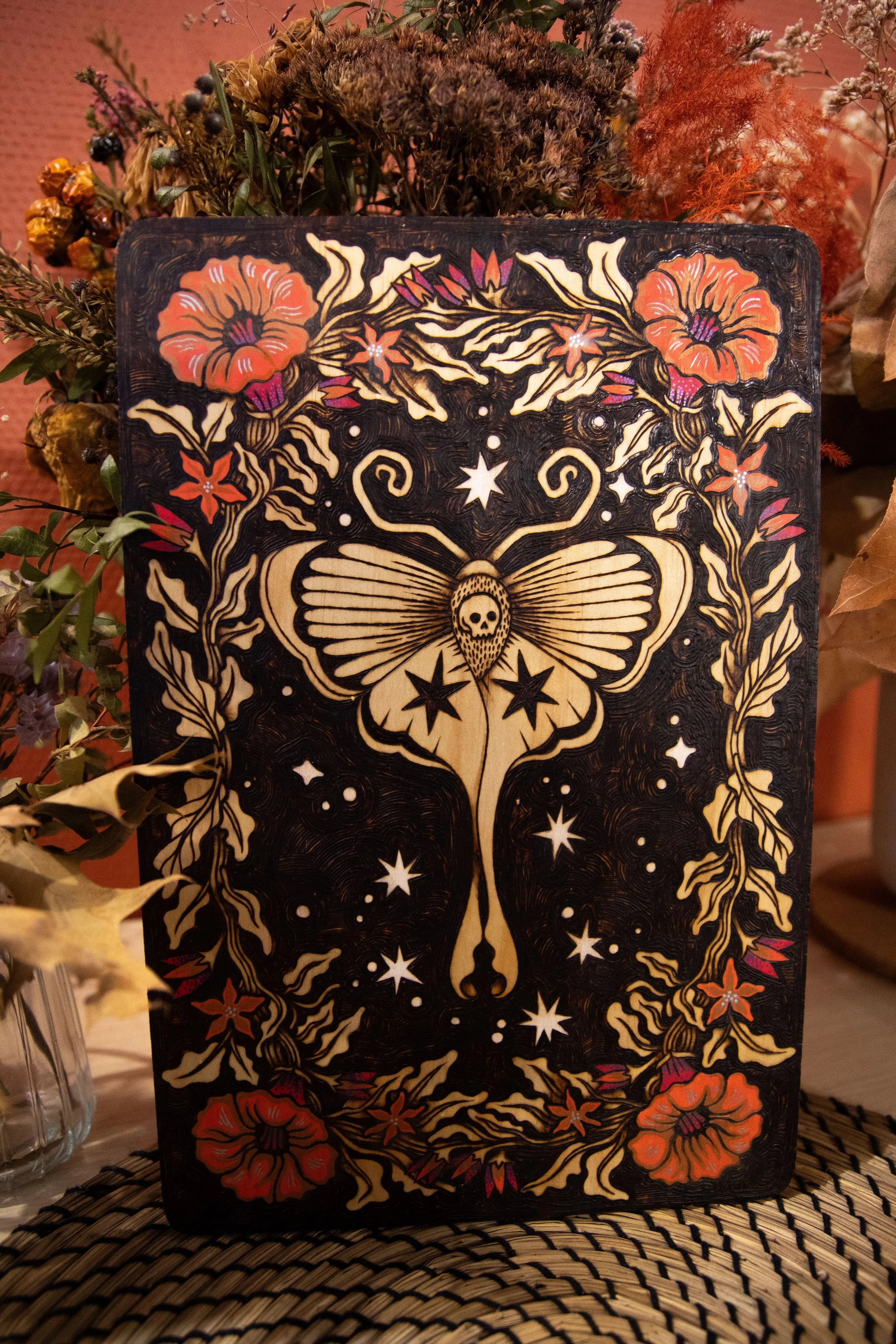 Plaque en bois artisanale pyrogravée d'un papillon de nuit mystique entouré de fleurs délicates sublimées par des touches de peinture colorée dans des teintes orangées.