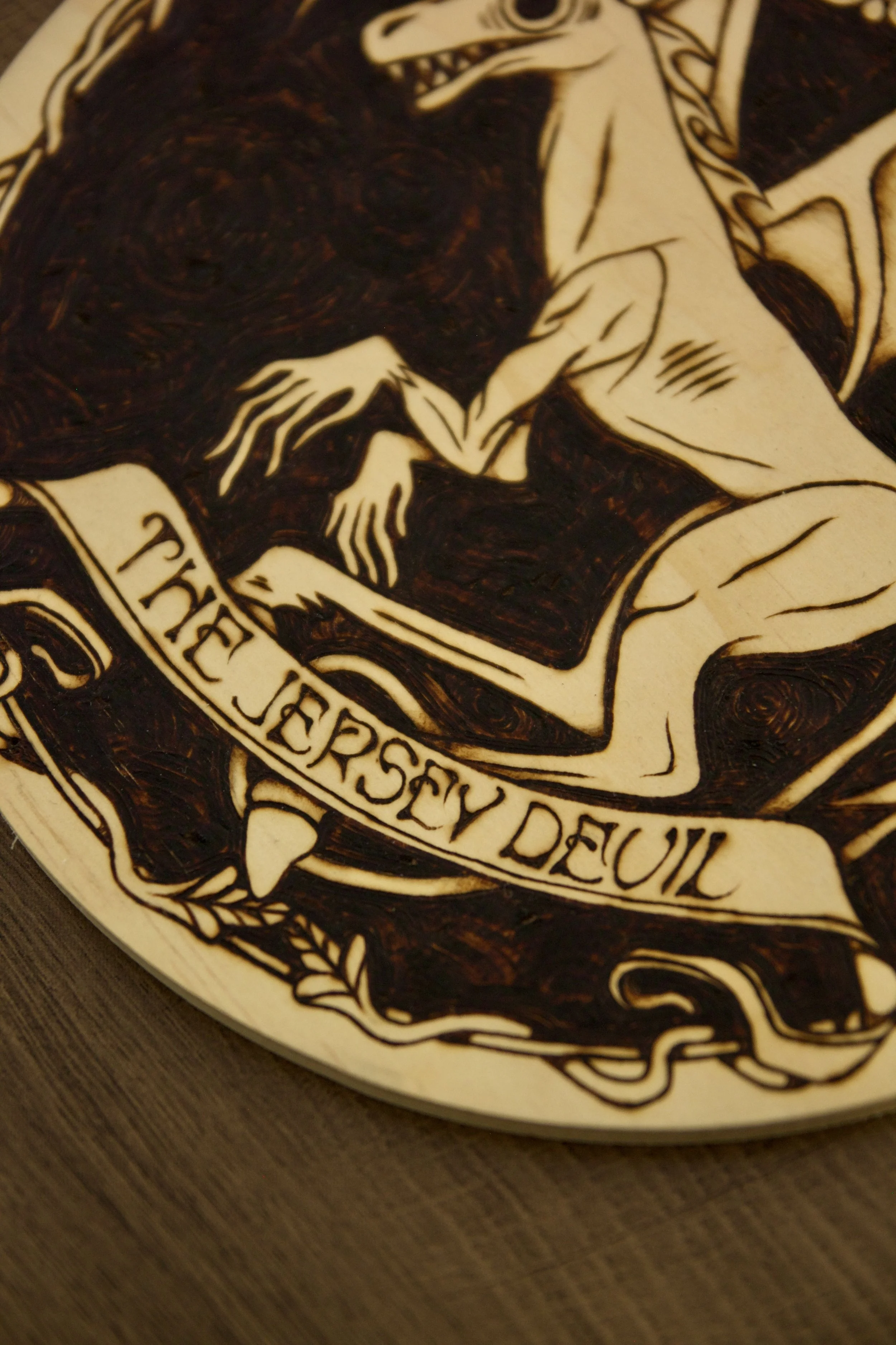 Détail de typographie d'une pyrogravure artisanale ronde de 20x20 cm sur bois représentant la silhouette du Diable de Jersey, créature ailée mythique du folklore américain.