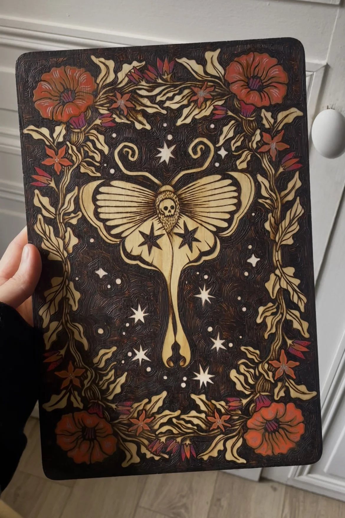 Plaque en bois artisanale pyrogravée d'un papillon de nuit mystique entouré de fleurs délicates sublimées par des touches de peinture colorée tenue en main.