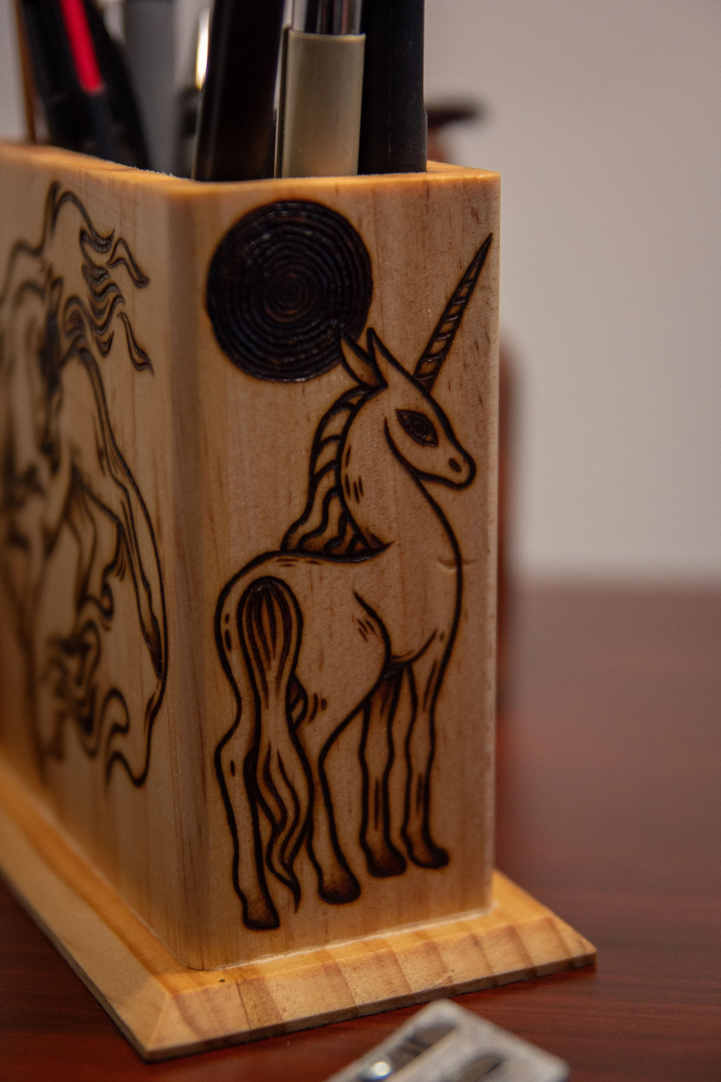 Vue de côté d'un pot à crayons carré en bois artisanal, montrant une face pyrogravée avec soin représentant une licorne.