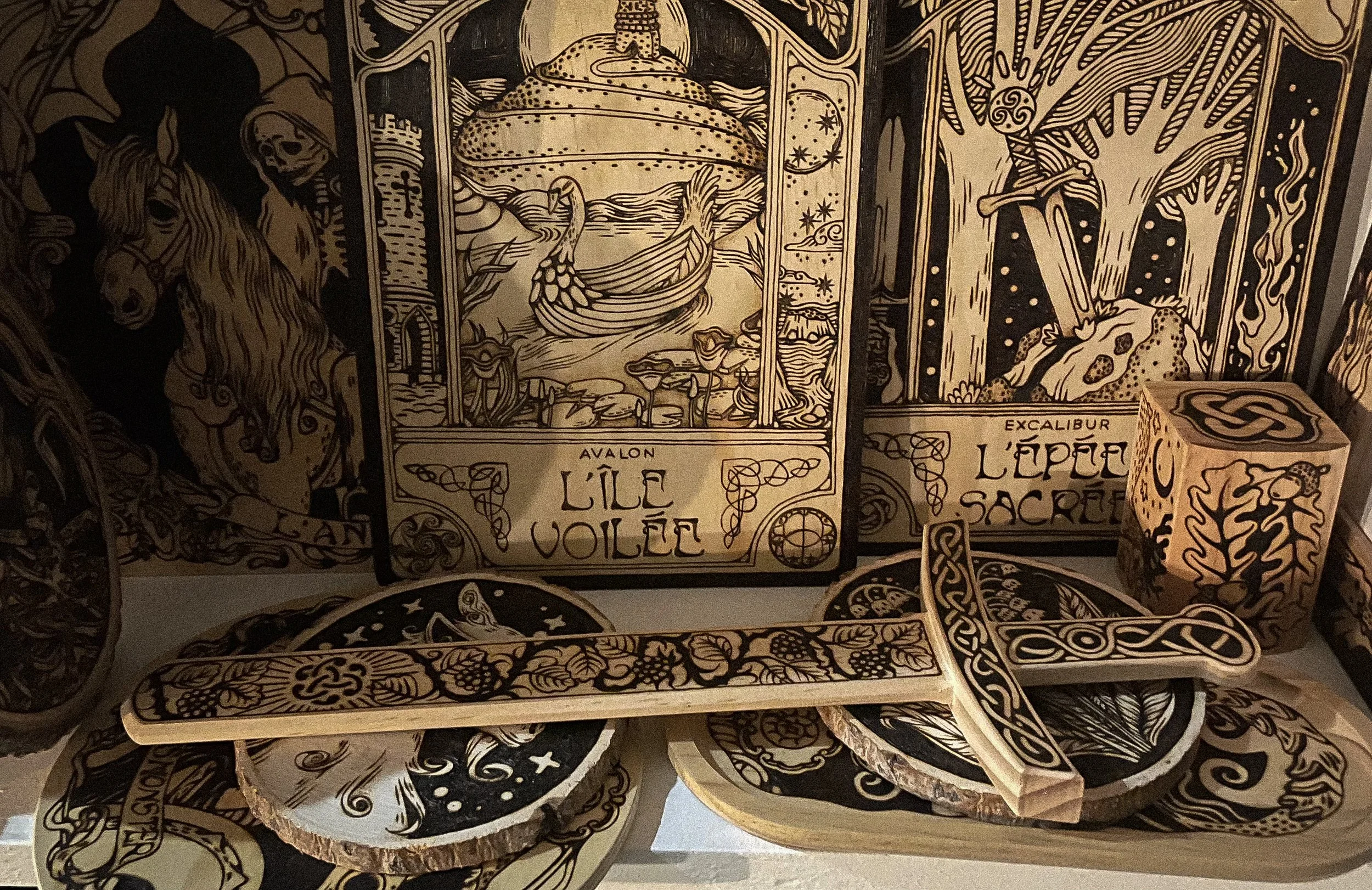 Étagère remplie d'objets en bois décorés avec des motifs celtique et médiévaux, incluant une épée, des encadrements, une boîte à bijoux, et des panneaux illustrés représentant des thèmes inspirés du folklore et du fantastique.