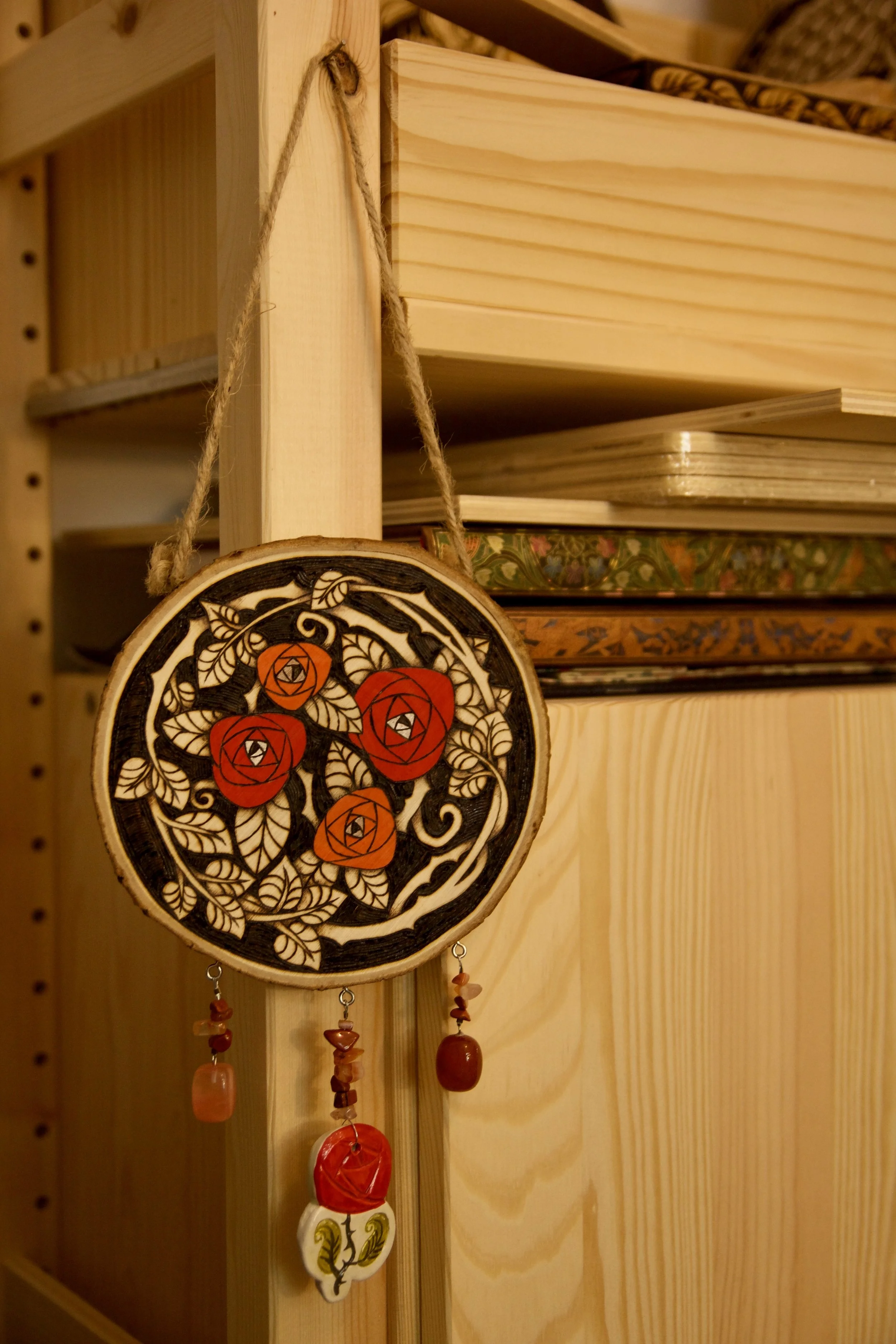 Amulette murale ronde en bois pyrogravé et peint avec des roses rouges, ornée de pendants en pierre et d'une rose en céramique.